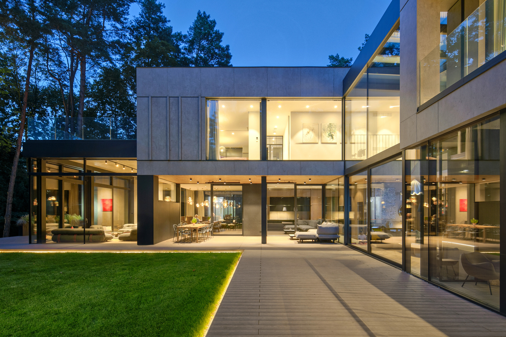 Lit living spaces opening onto a plank terrace and lawn edged with a glowing strip at dusk