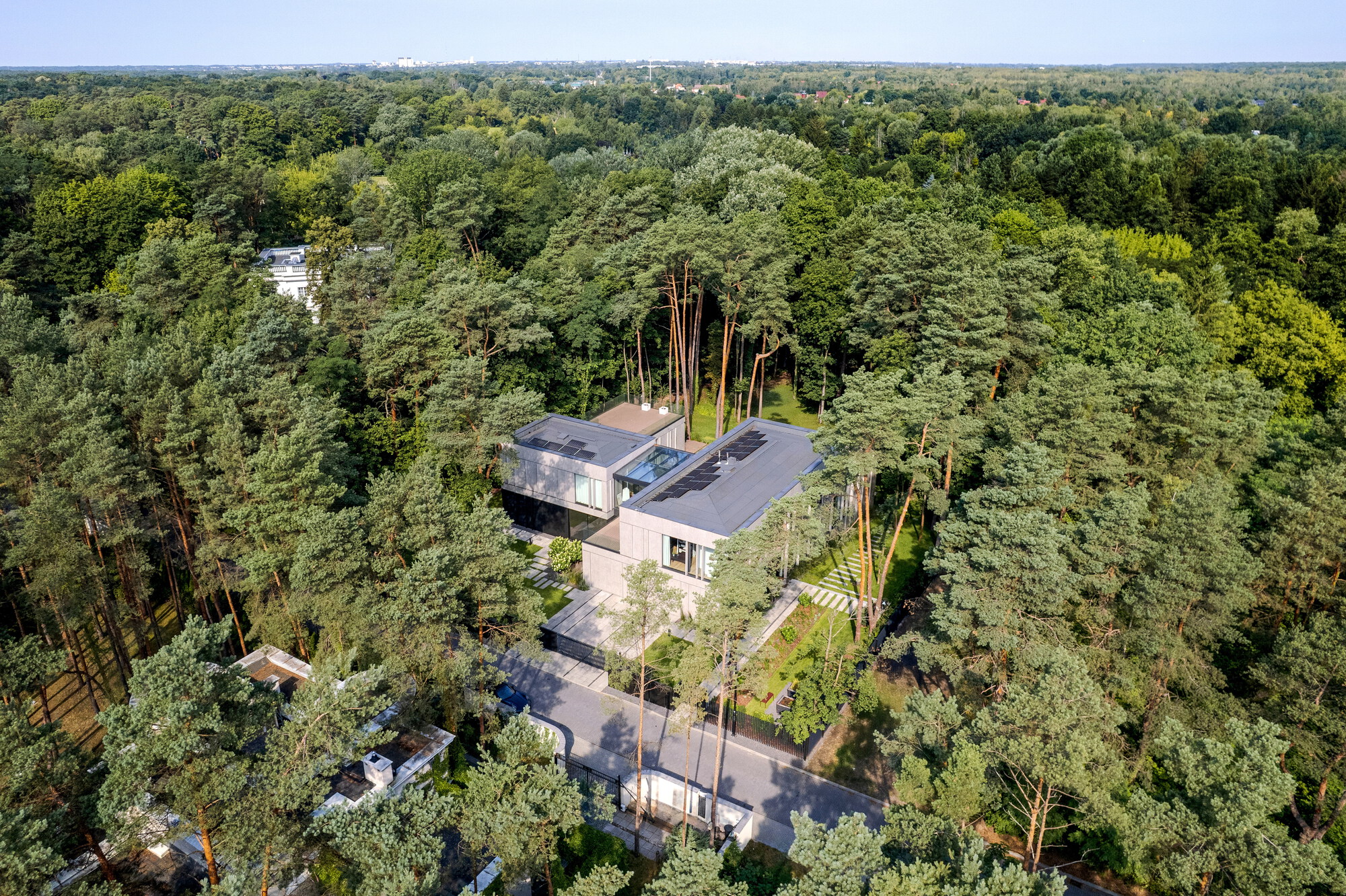 Aerial view of the flat-roofed house with solar panels set among dense pine forest beside a quiet road