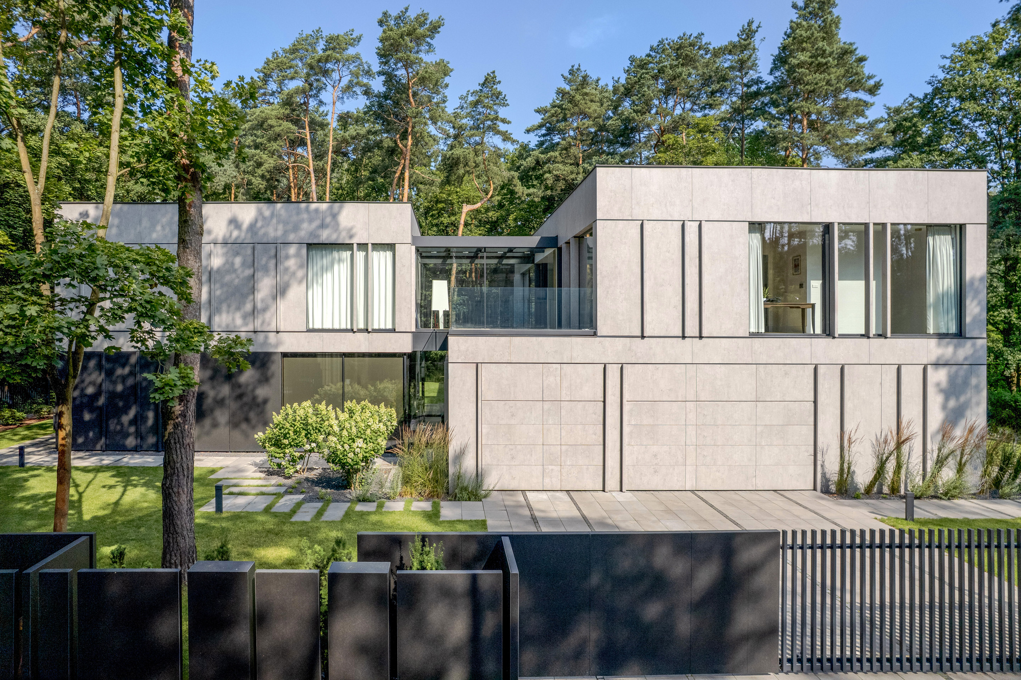 Street facade of two stacked grey panel volumes with a glass-balustraded terrace, framed by pine trees in summer sun