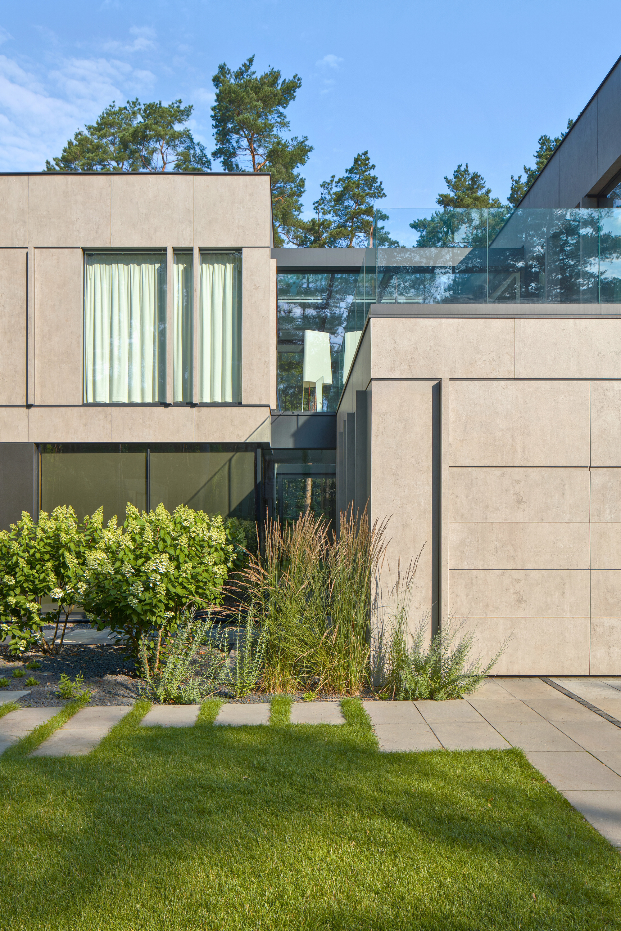 Detail of the panel facade and a glass-railed link, with hydrangeas and grasses in a gravel bed beside a lawn path