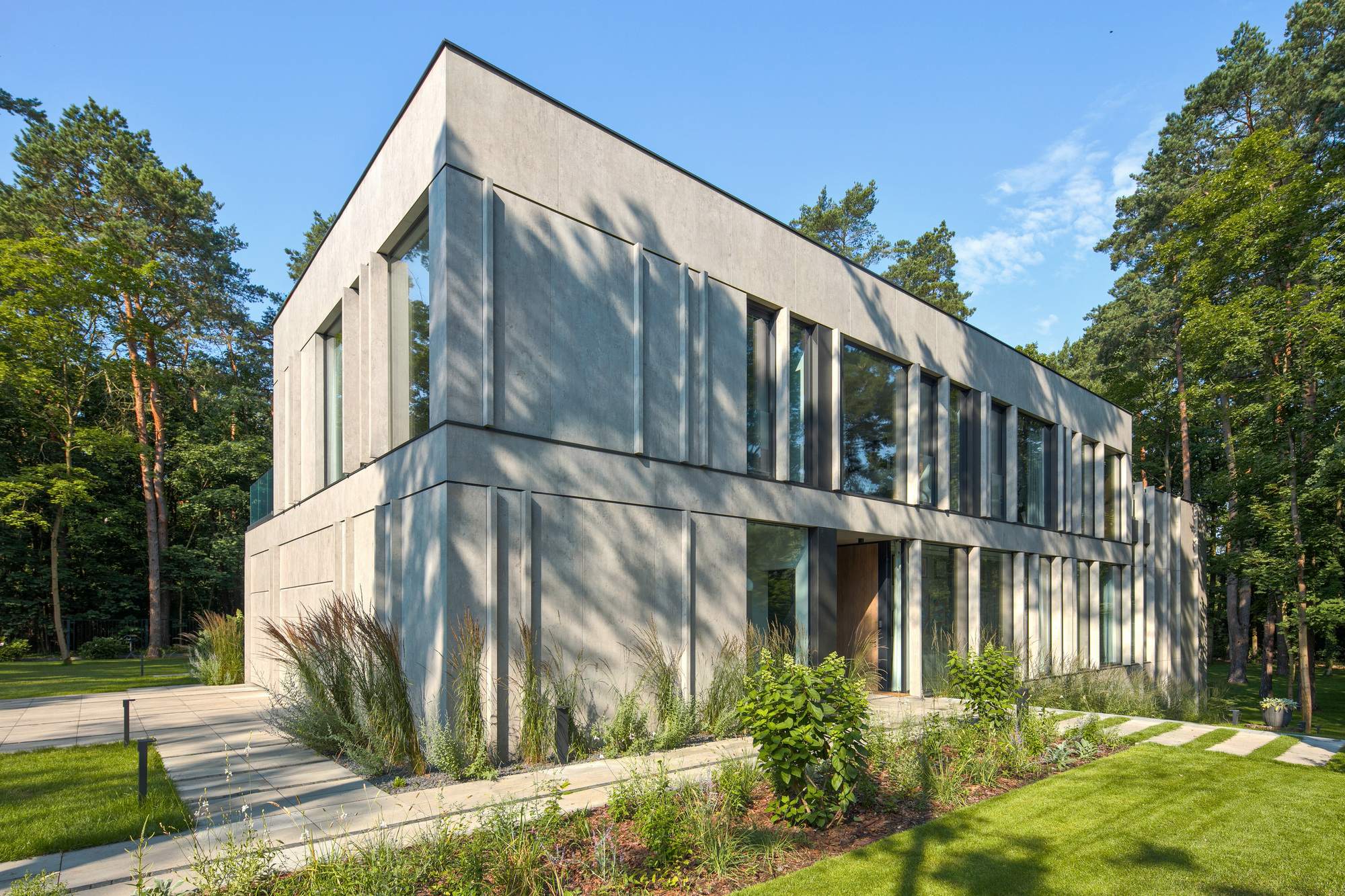 Corner of the two-storey grey volume with tall vertical fins and glazing, set among pines and a planted border in sunlight