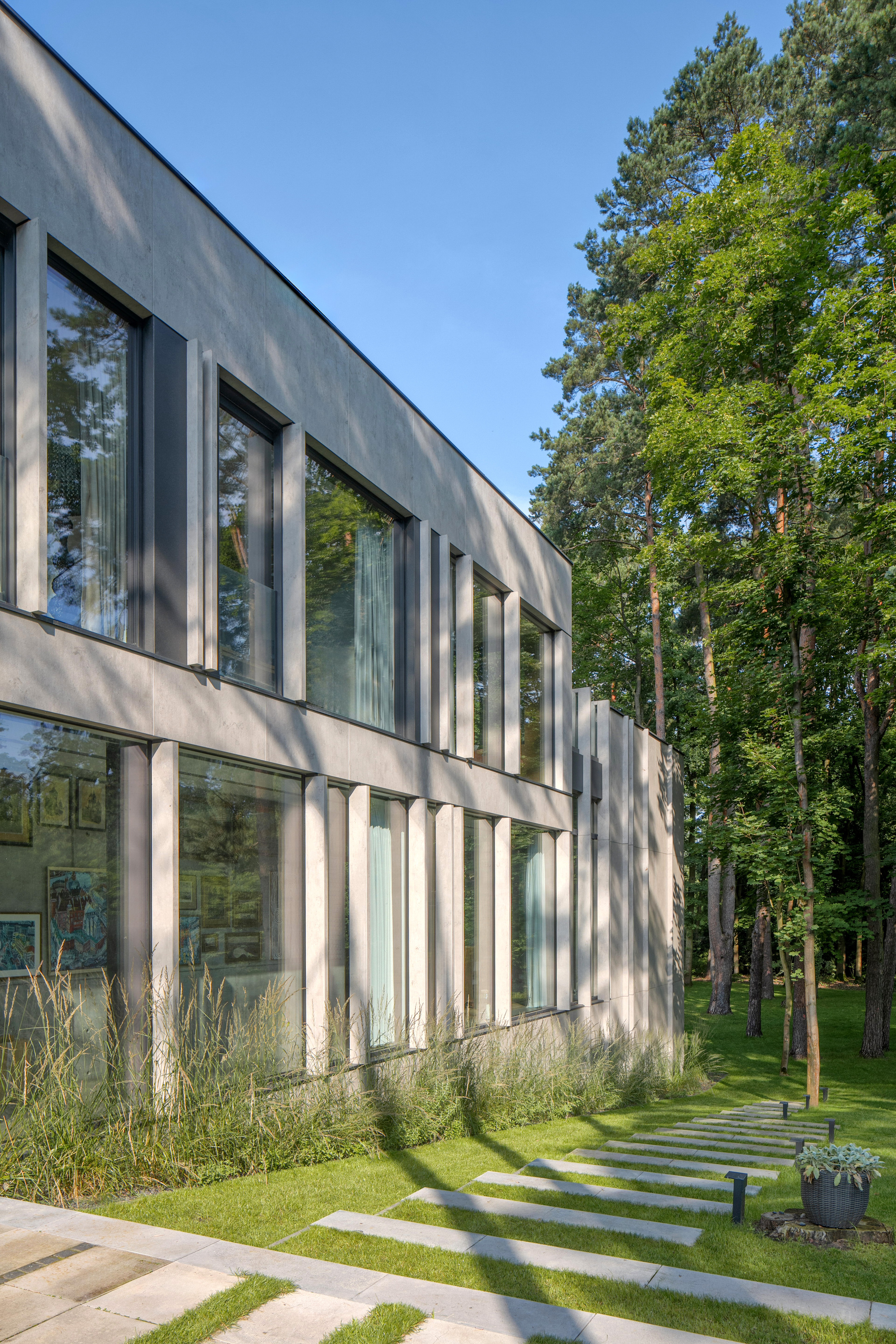 Side elevation with deep vertical fins between windows, stepping stones crossing the lawn under tall trees in late afternoon light