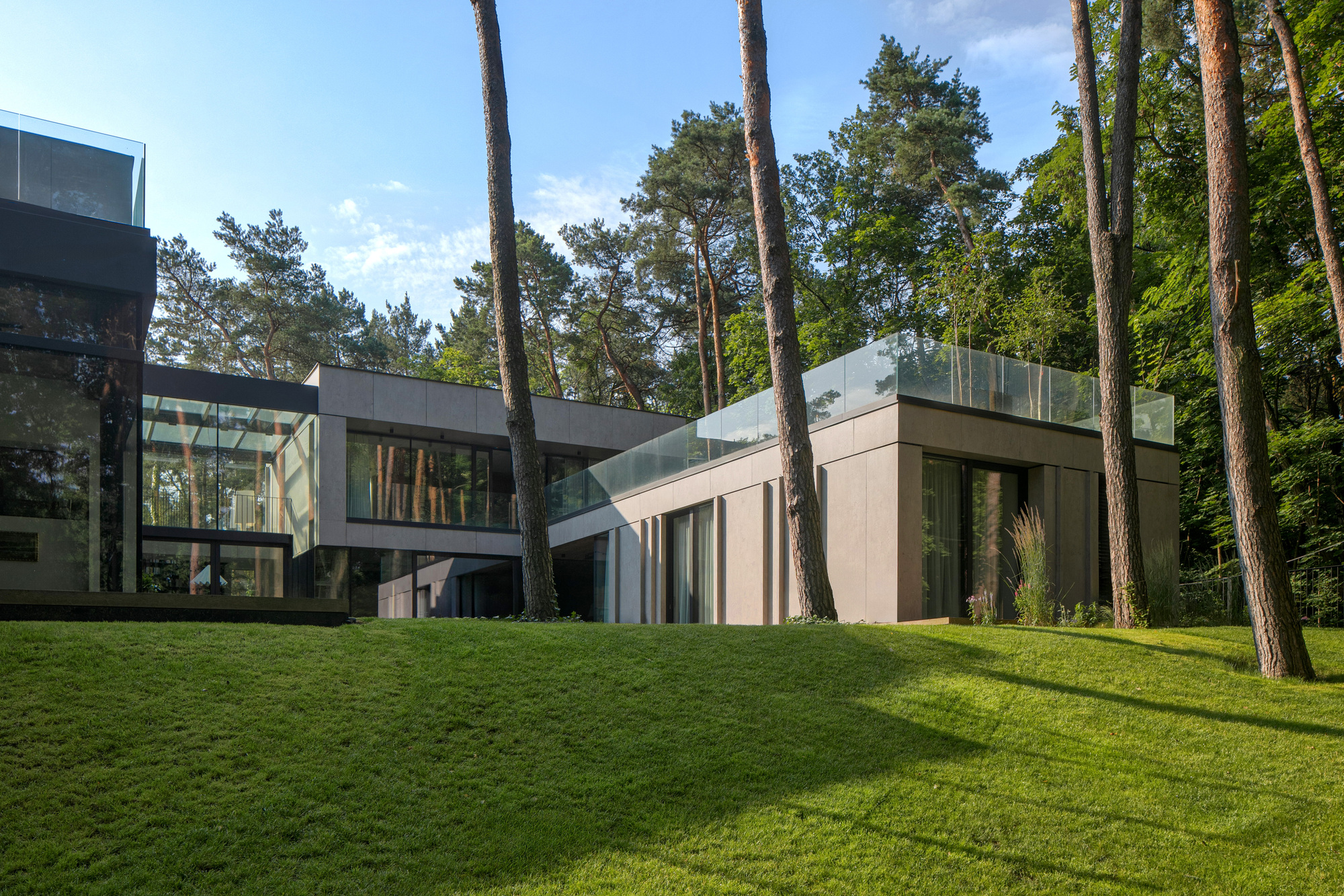 Two-storey house of concrete panels and glass with a rooftop glass balustrade, set among tall pines above a sunlit grass slope