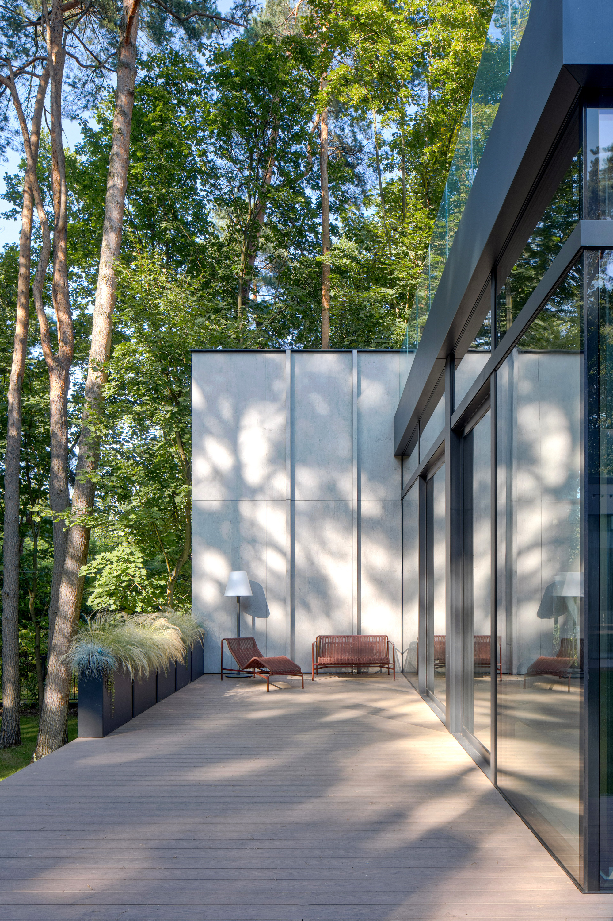 Timber deck terrace with red lounge chair and bench against a pale panel wall, dappled by pine shadows