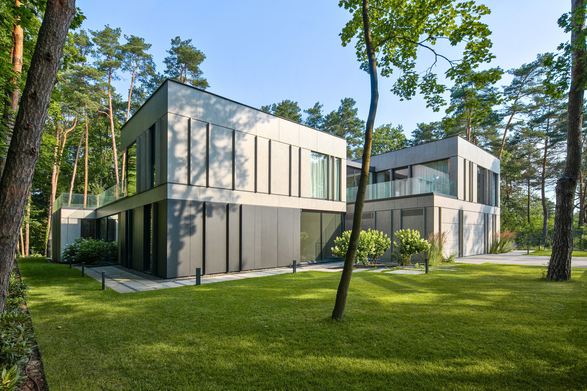 Corner view of two panelled volumes with vertical dark joints, a paved path and a thin tree trunk on the lawn