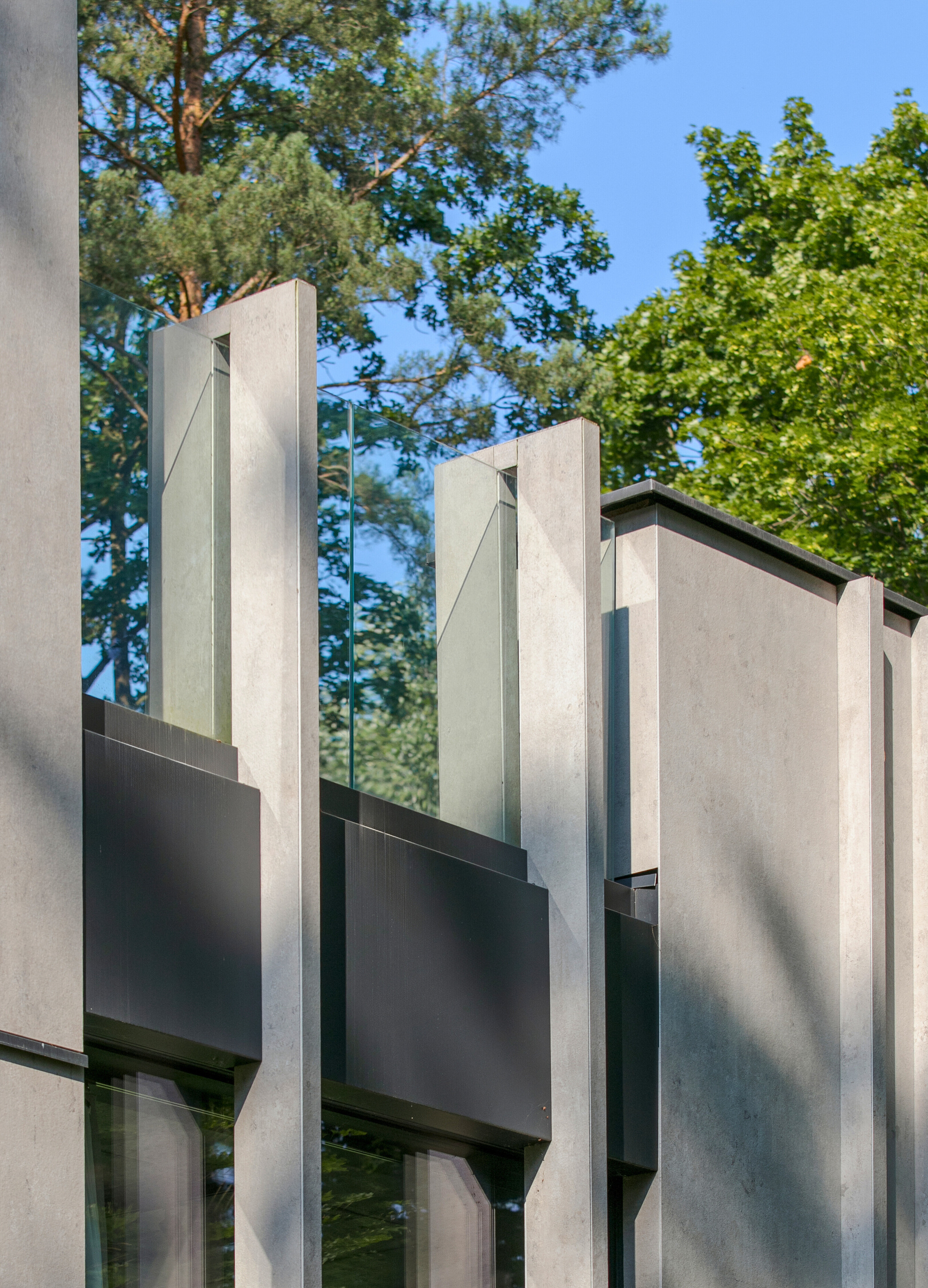 Close-up of concrete fins and dark metal spandrel panels with glass parapet, framed by pine and leafy branches in summer sun