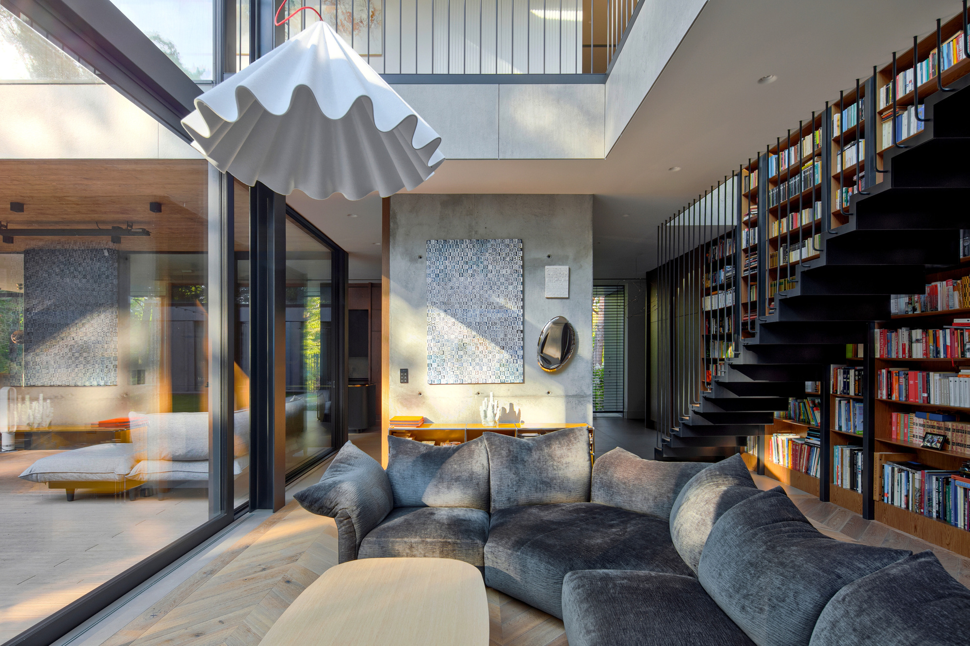 Double-height living room with grey sofa, pleated white pendant lamp and black steel stair beside a built-in bookcase wall