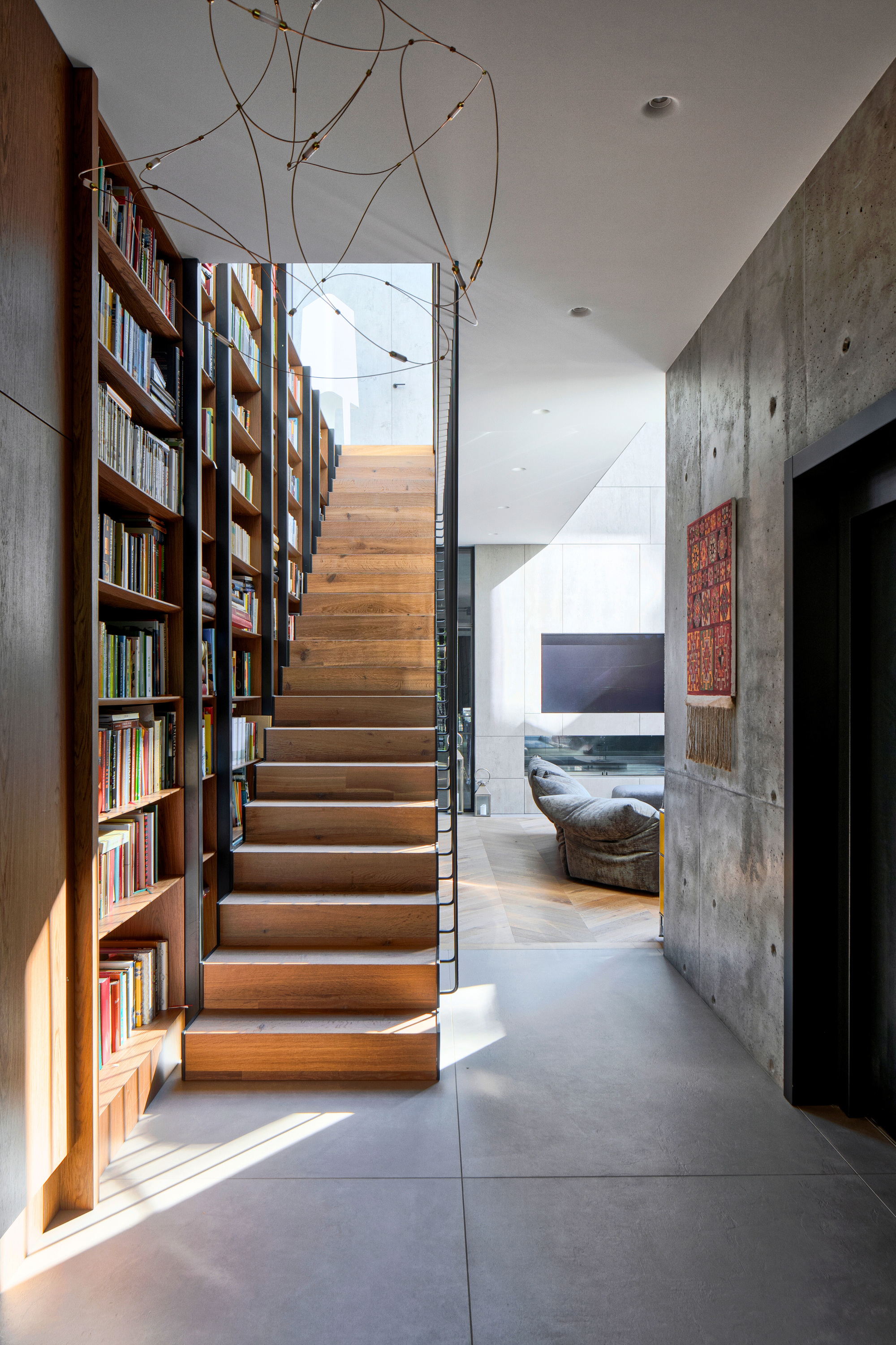 Timber stair lined with a full-height bookcase, leading toward a board-formed concrete wall in a streak of afternoon sunlight