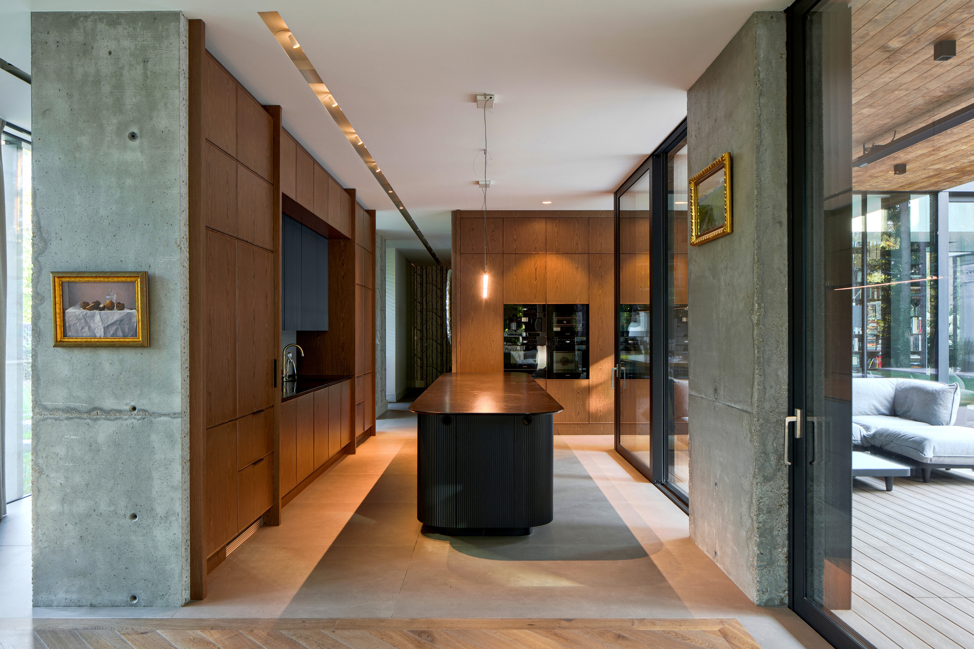 Kitchen with walnut-toned cabinetry and dark island between raw concrete piers, with framed paintings and a glazed door opening to the deck