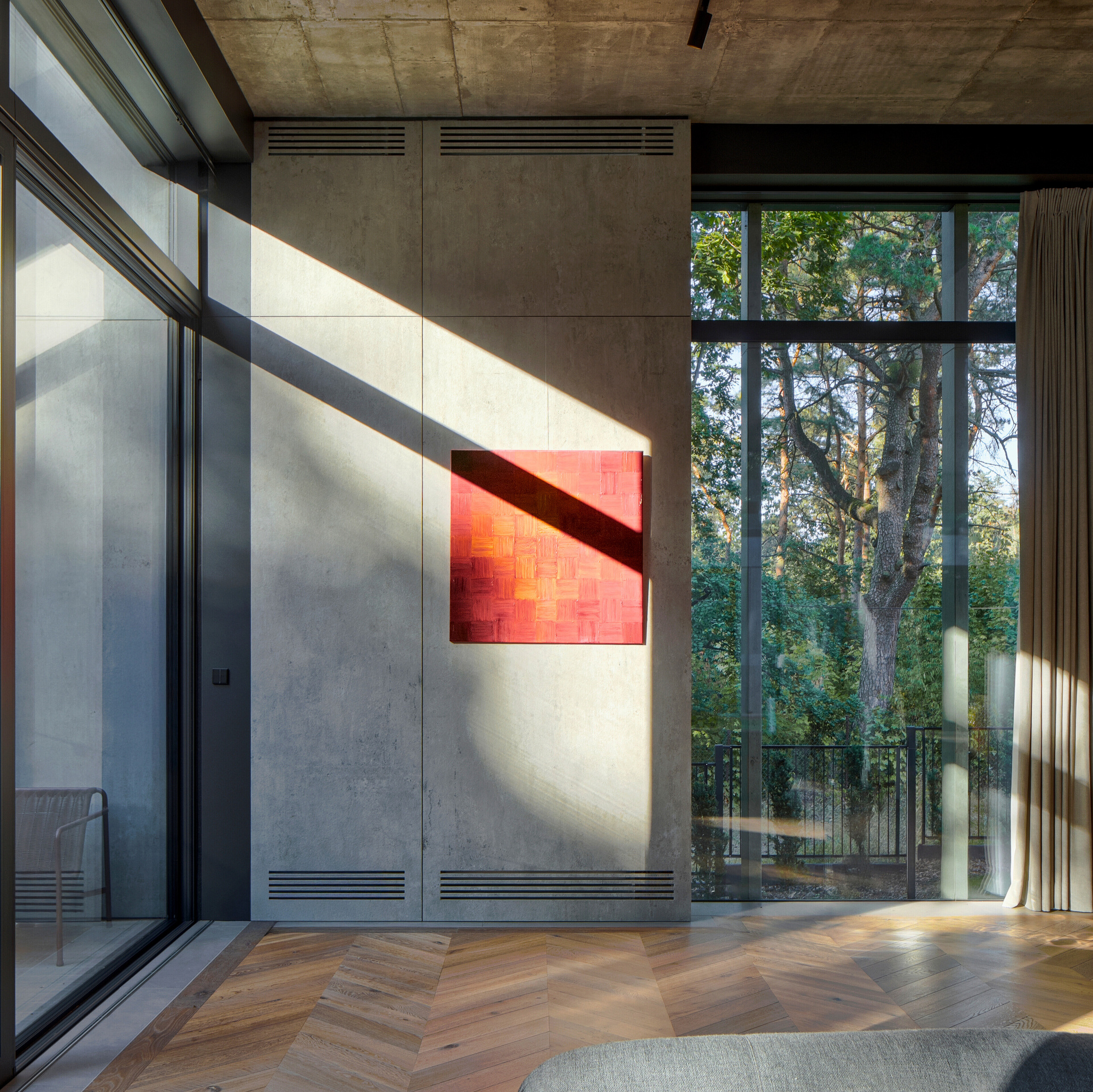 Concrete panel wall with red square painting crossed by a diagonal shadow, beside floor-to-ceiling glass looking onto pine trunks
