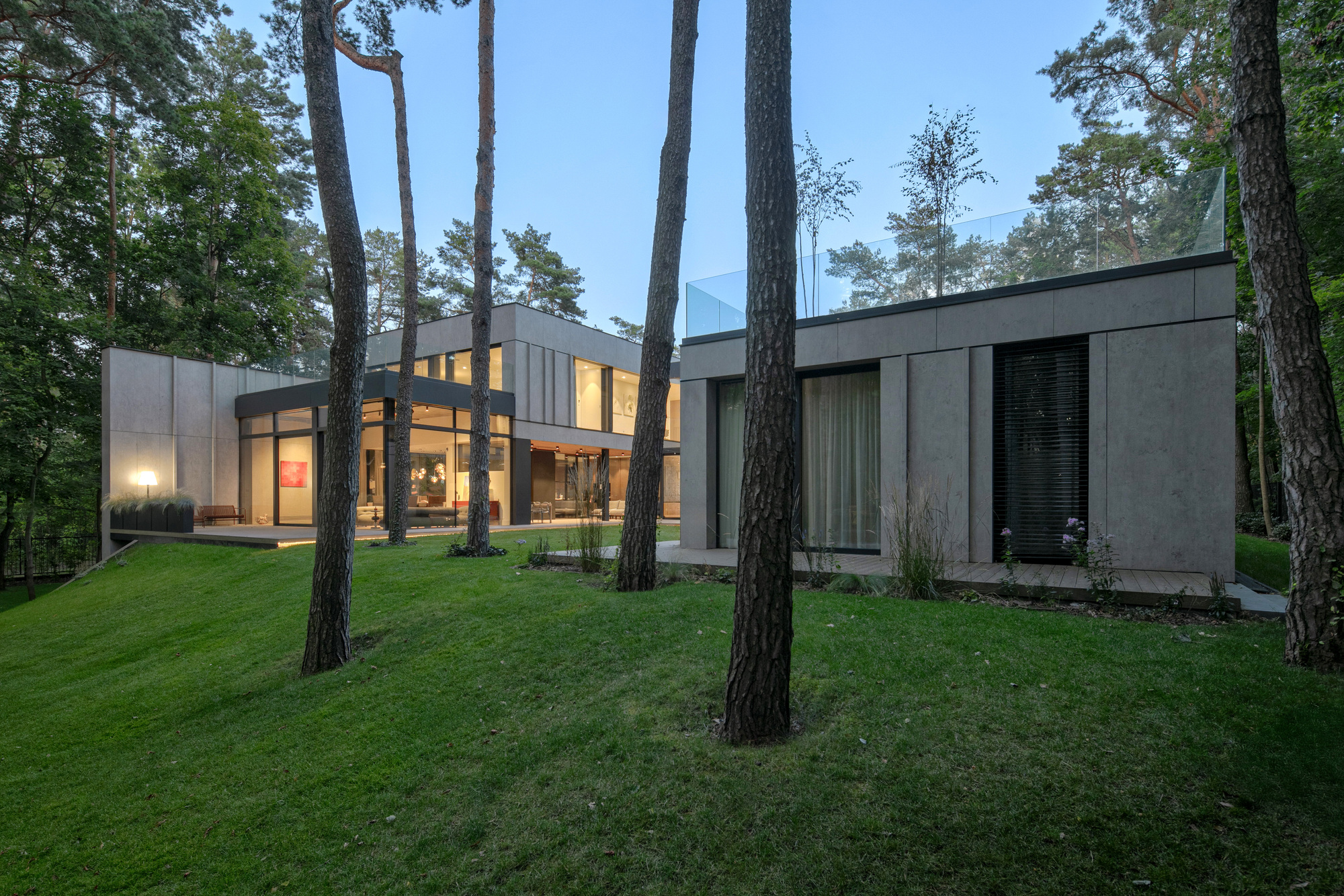 Concrete-clad volumes with glass parapet glowing at dusk, seen across a grassy slope between tall pine trunks