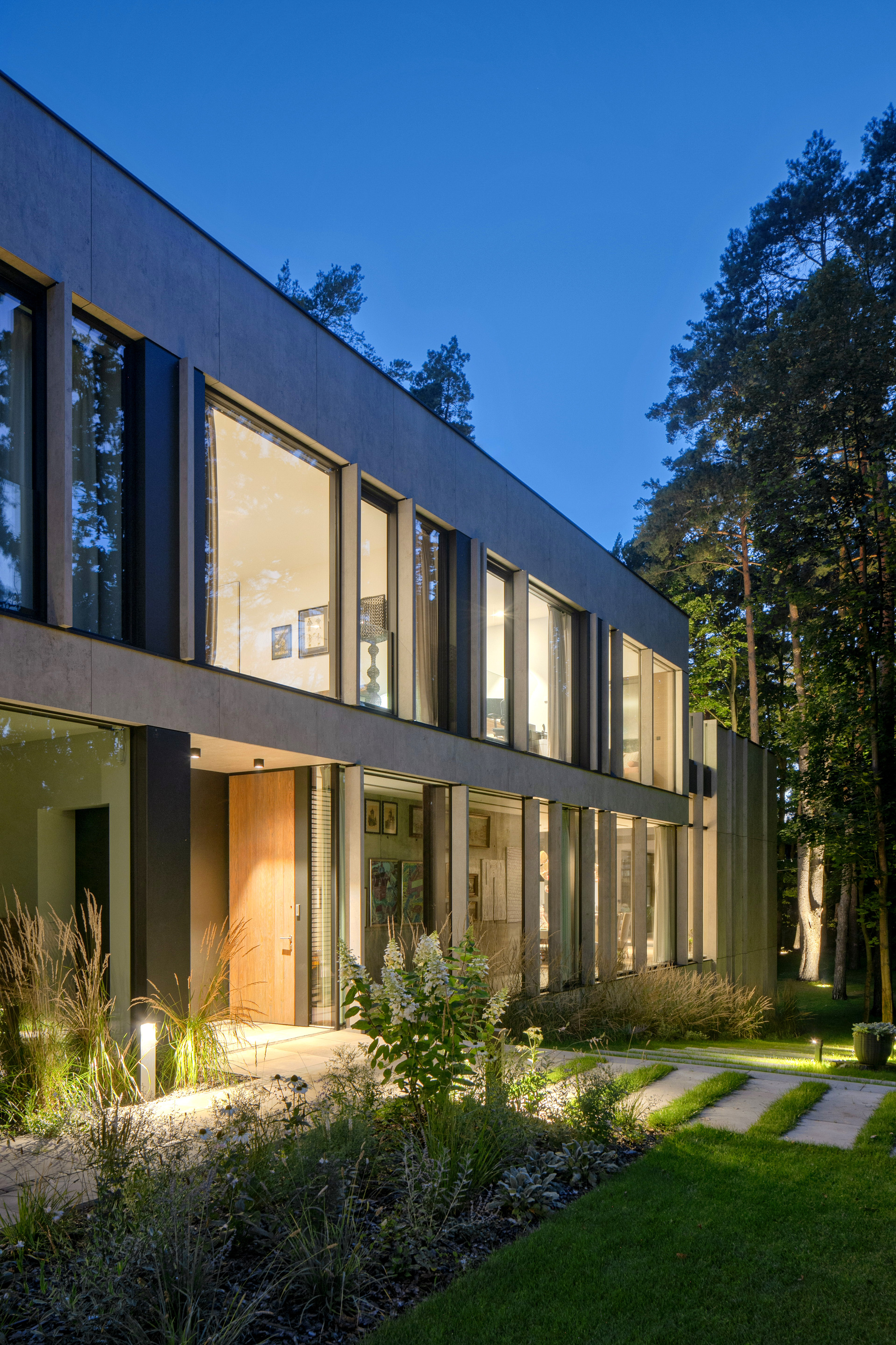 Two-storey facade of grey panels and tall glazing with a timber entrance door, lit at dusk beside pines and grass beds