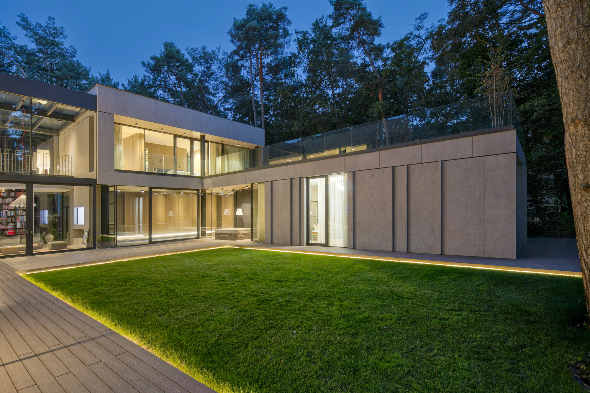 Courtyard lawn edged with glowing strip lights, framed by glazed walls and panelled facade under tall pines at dusk