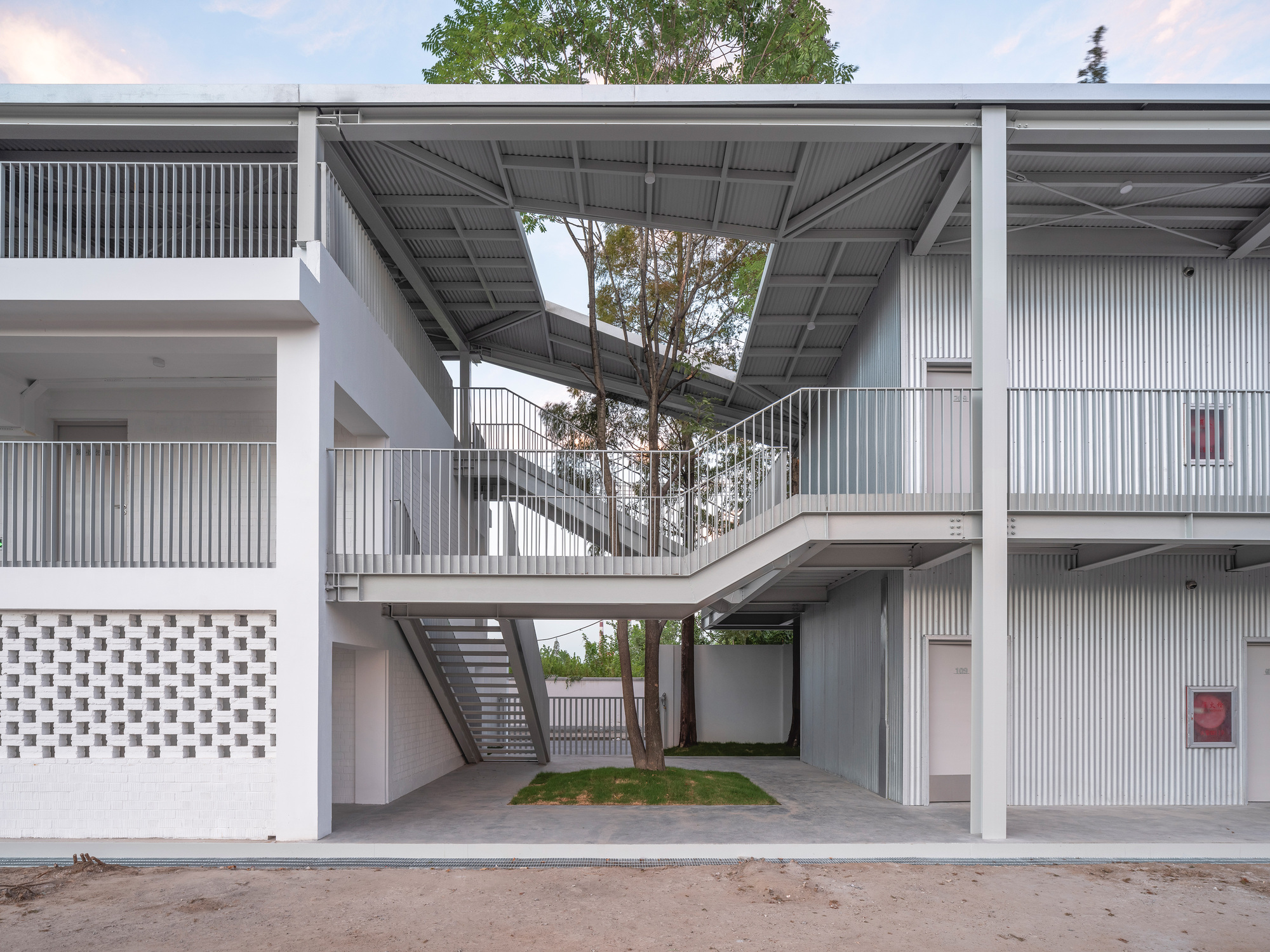 Two-storey courtyard with a steel stair and walkway beneath a sloping roof, a tree rising through the opening in soft evening light
