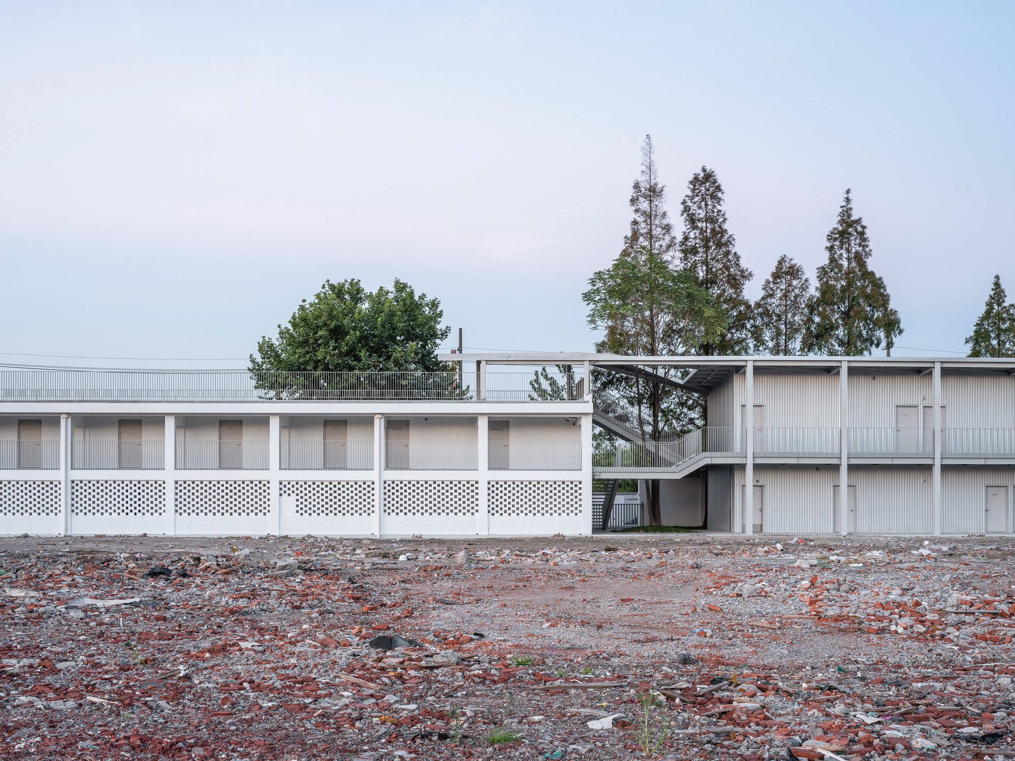 Long white two-storey block with perforated brick base seen across a rubble-strewn lot, tall conifers behind under a pale dusk sky