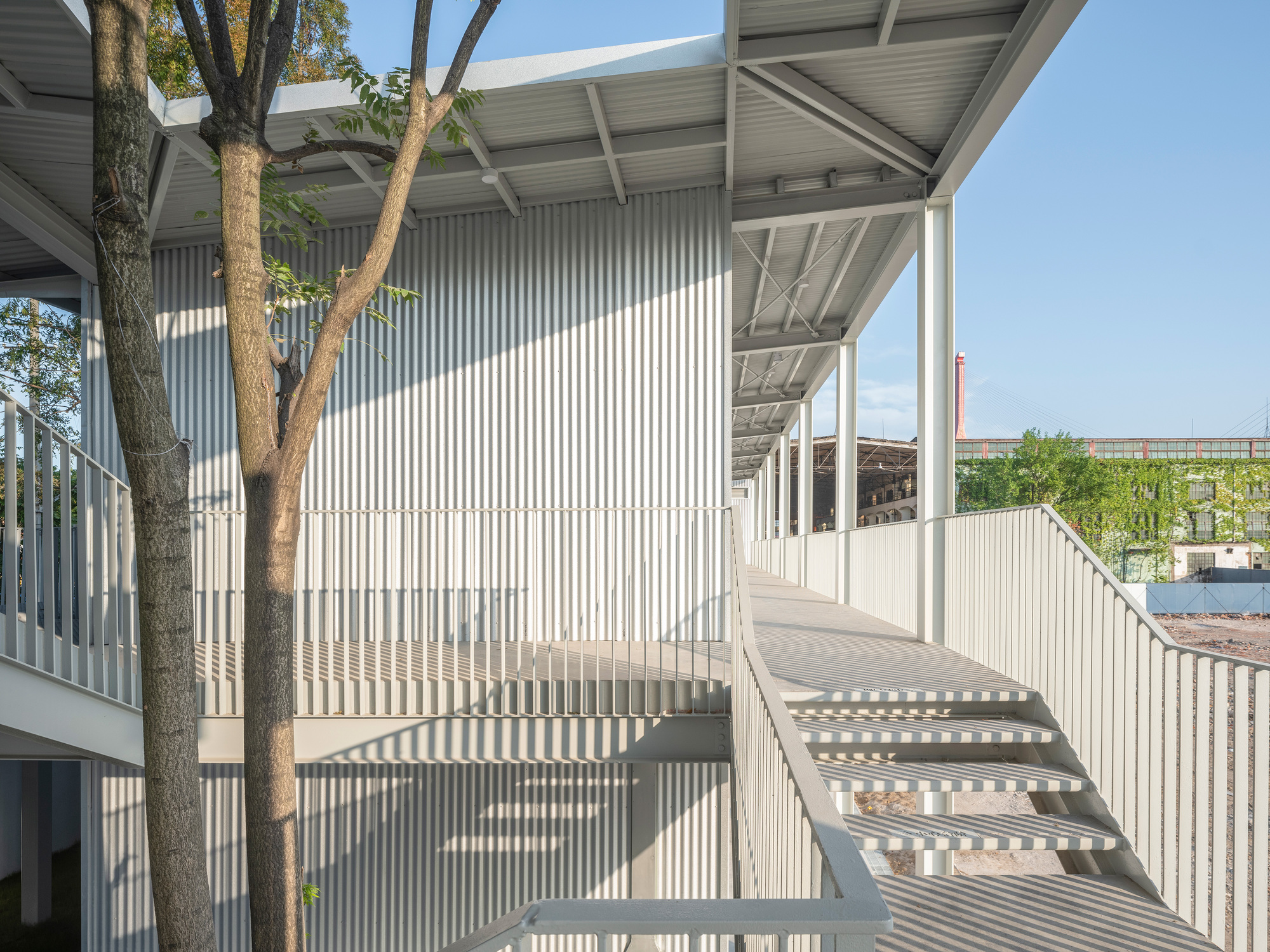 Steel stair and slatted railings beside corrugated metal walls, with tree trunks in the foreground and strong sunlight casting stripes