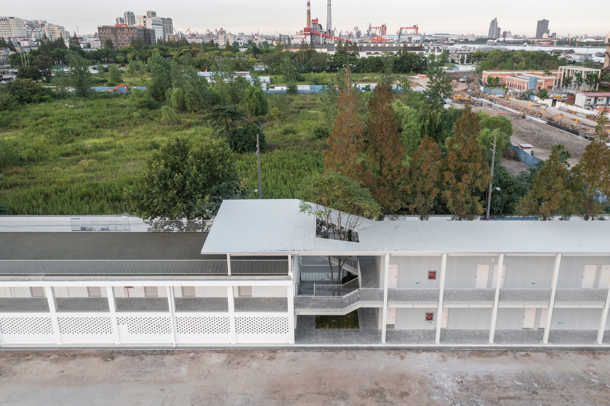 Aerial view of the white-roofed two-storey building with a tree through the roof opening, overlooking wetland grass and a distant industrial skyline