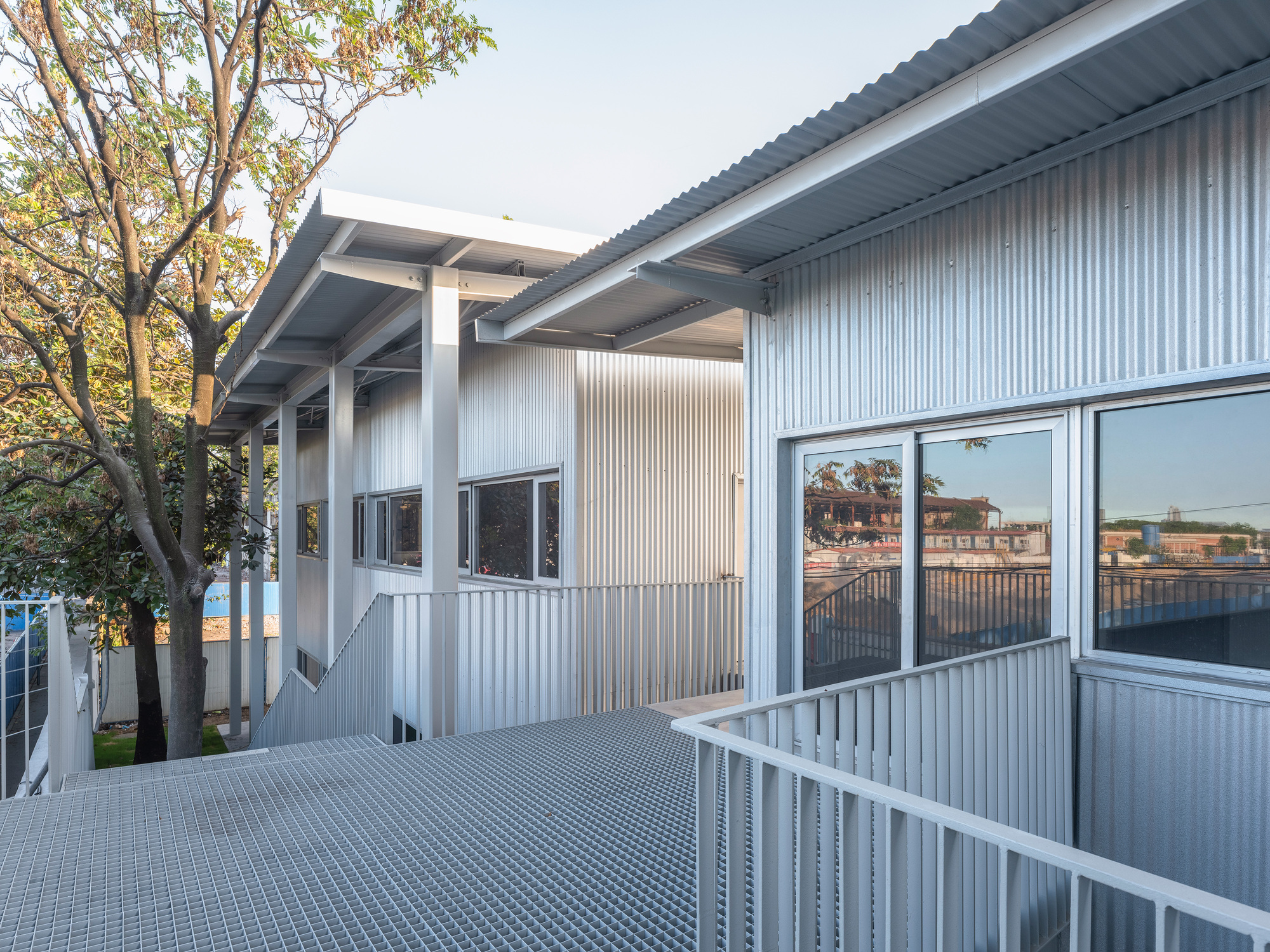 Grated steel walkway beside corrugated metal walls and windows reflecting the neighborhood, with a leafy tree at left in afternoon sun