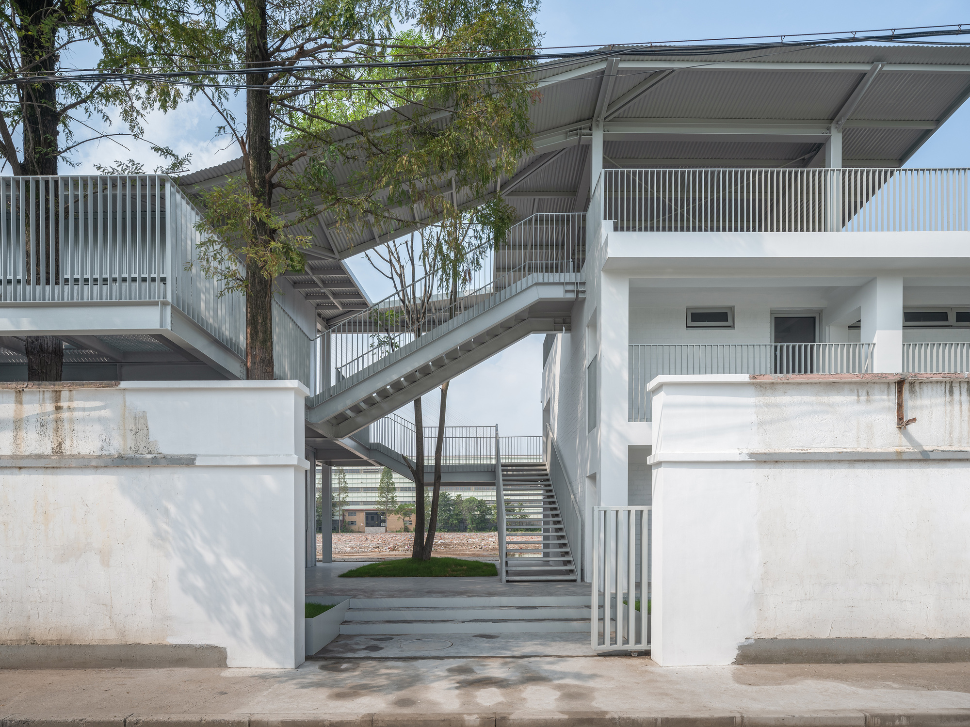 Street-side view of a steel staircase rising between whitewashed walls, with a tree growing through the upper walkway in hazy daylight
