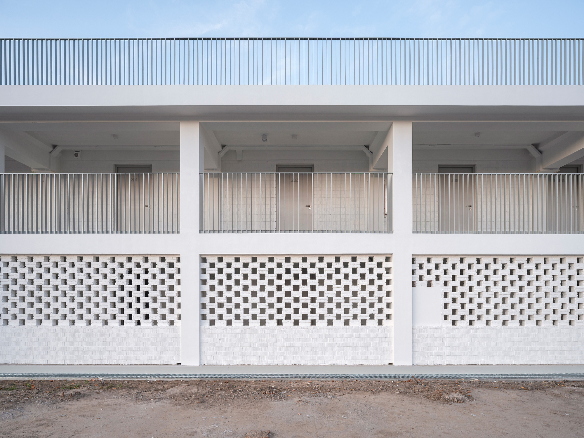 Two-storey white facade with a perforated brick screen below and a railed gallery above, seen across bare dirt at dusk