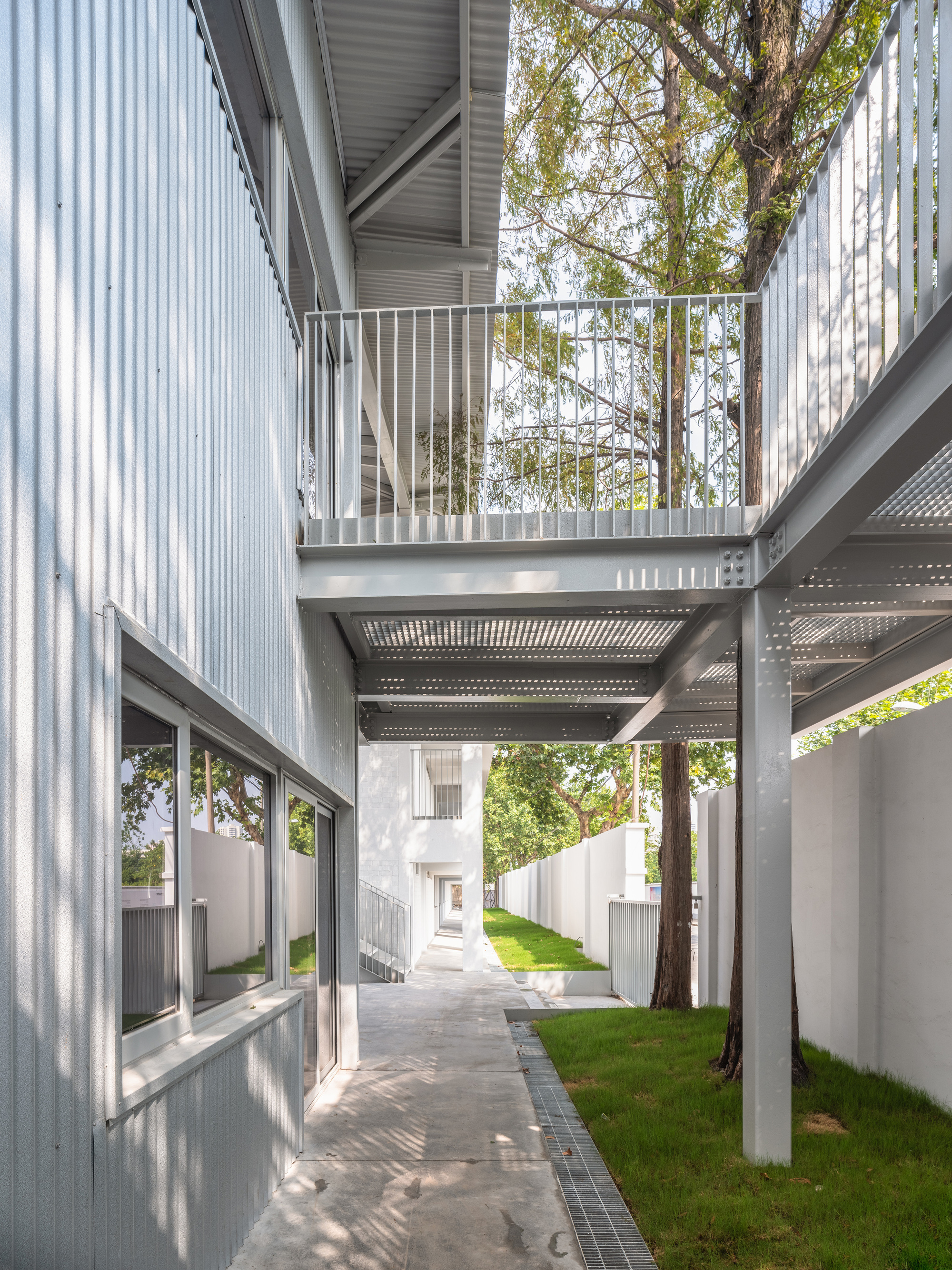 Narrow passage between corrugated metal wall and white boundary wall, with an elevated steel walkway crossing over a tree trunk