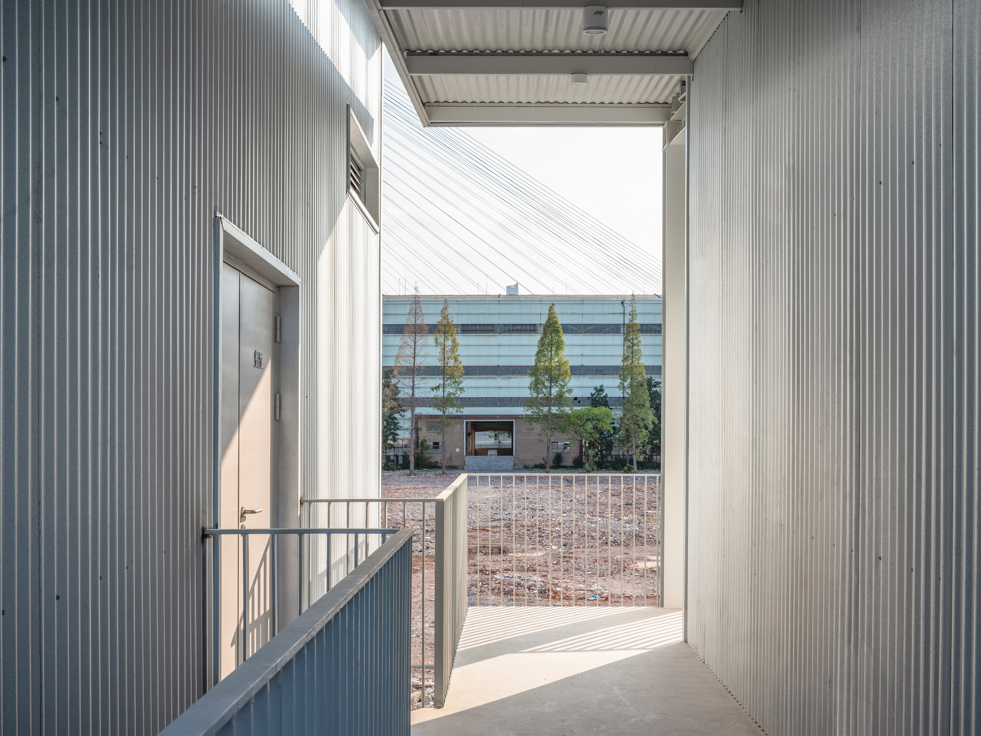 Corrugated metal corridor with a grey door and railing opening onto a cable-stayed bridge and factory building beyond bare ground