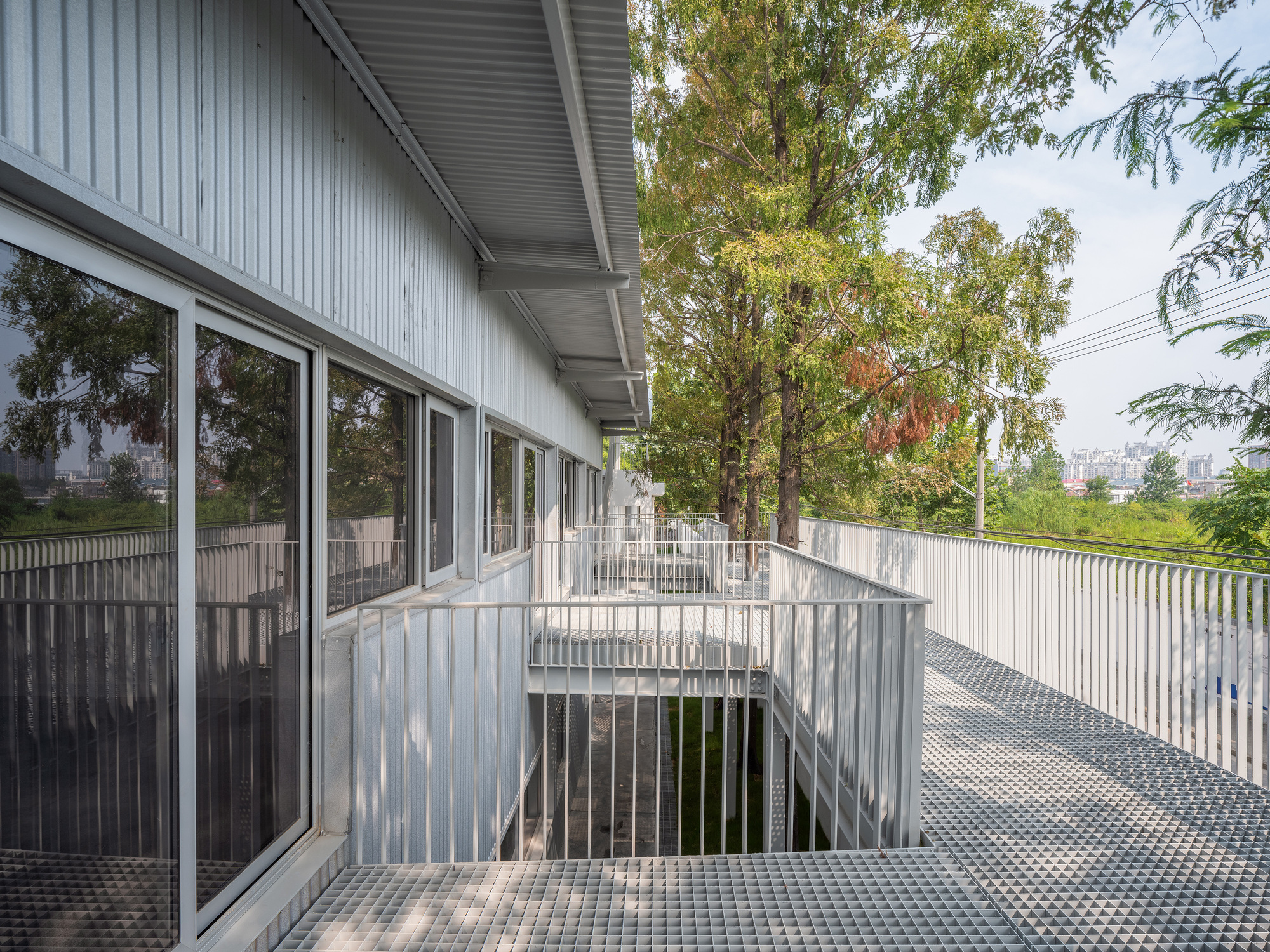 Upper walkway with square grating floor and slim vertical railings beside corrugated cladding, framed by autumn trees and distant city haze
