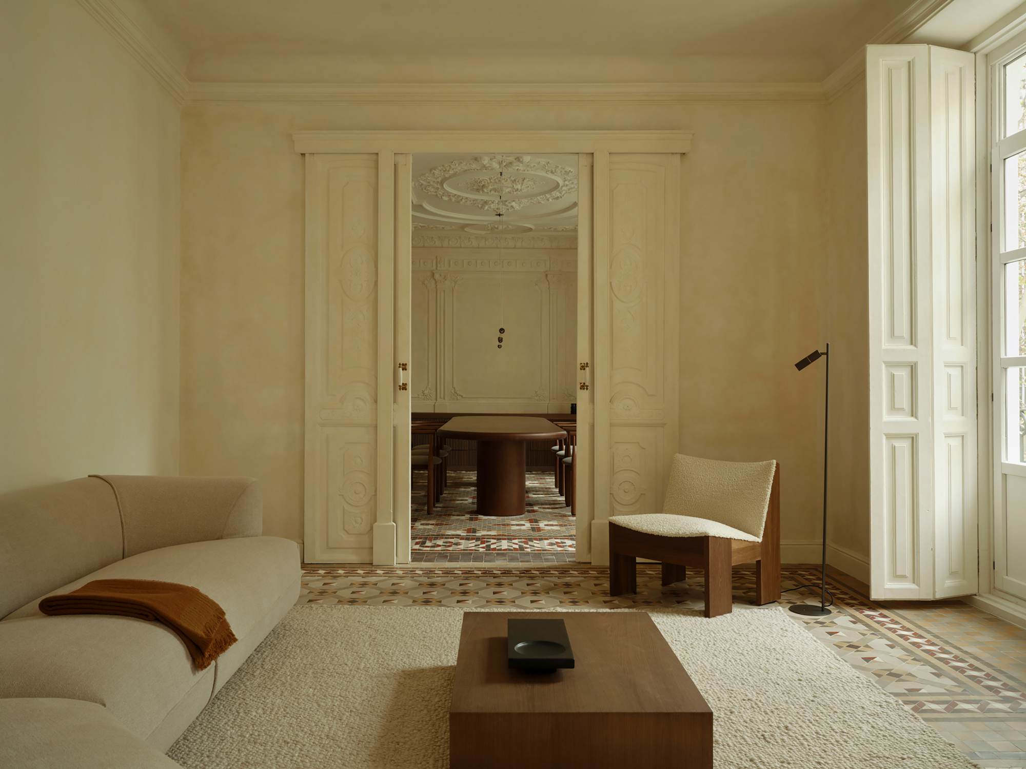 Living room with a curved cream sofa and walnut coffee table, looking through carved double doors to an oval dining table in soft daylight