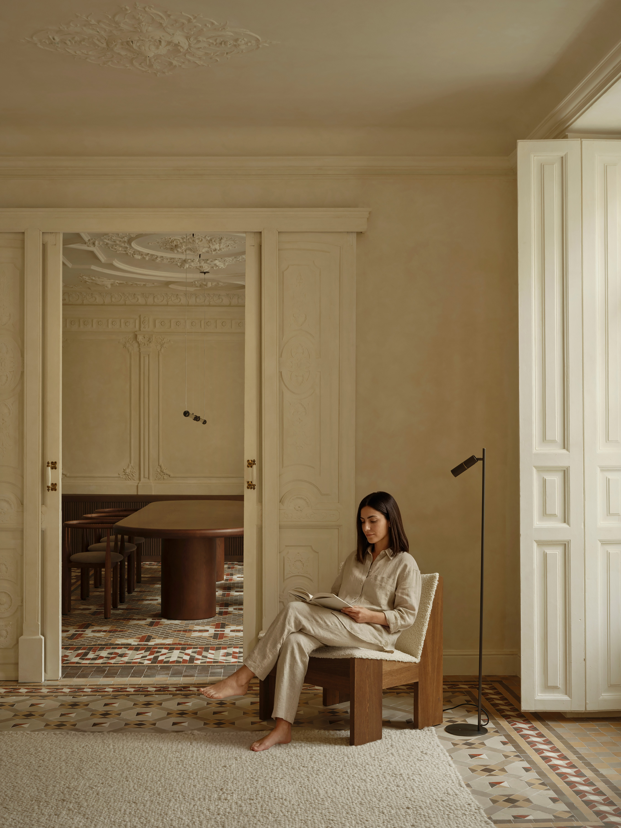Woman reading in a timber lounge chair beside tall white shutters, with the carved doorway and dining table behind her