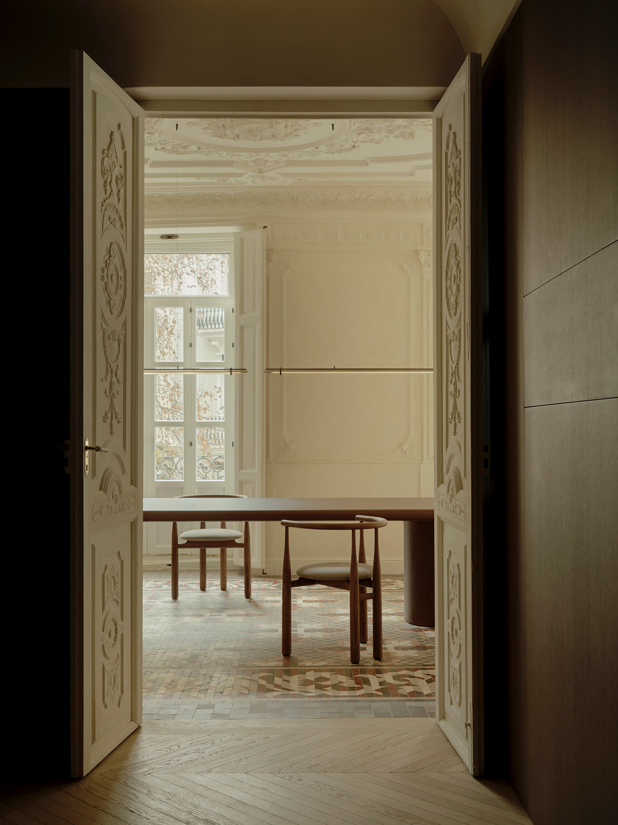 Open carved double doors frame a dining table and chairs against a tall window, with chevron parquet in the foreground