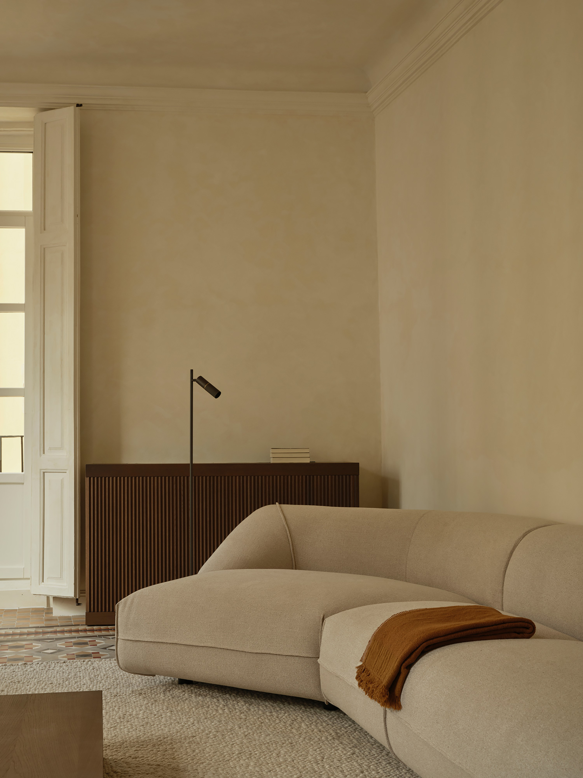 Curved beige sofa with a rust throw in front of a fluted timber sideboard and black floor lamp, window light at left