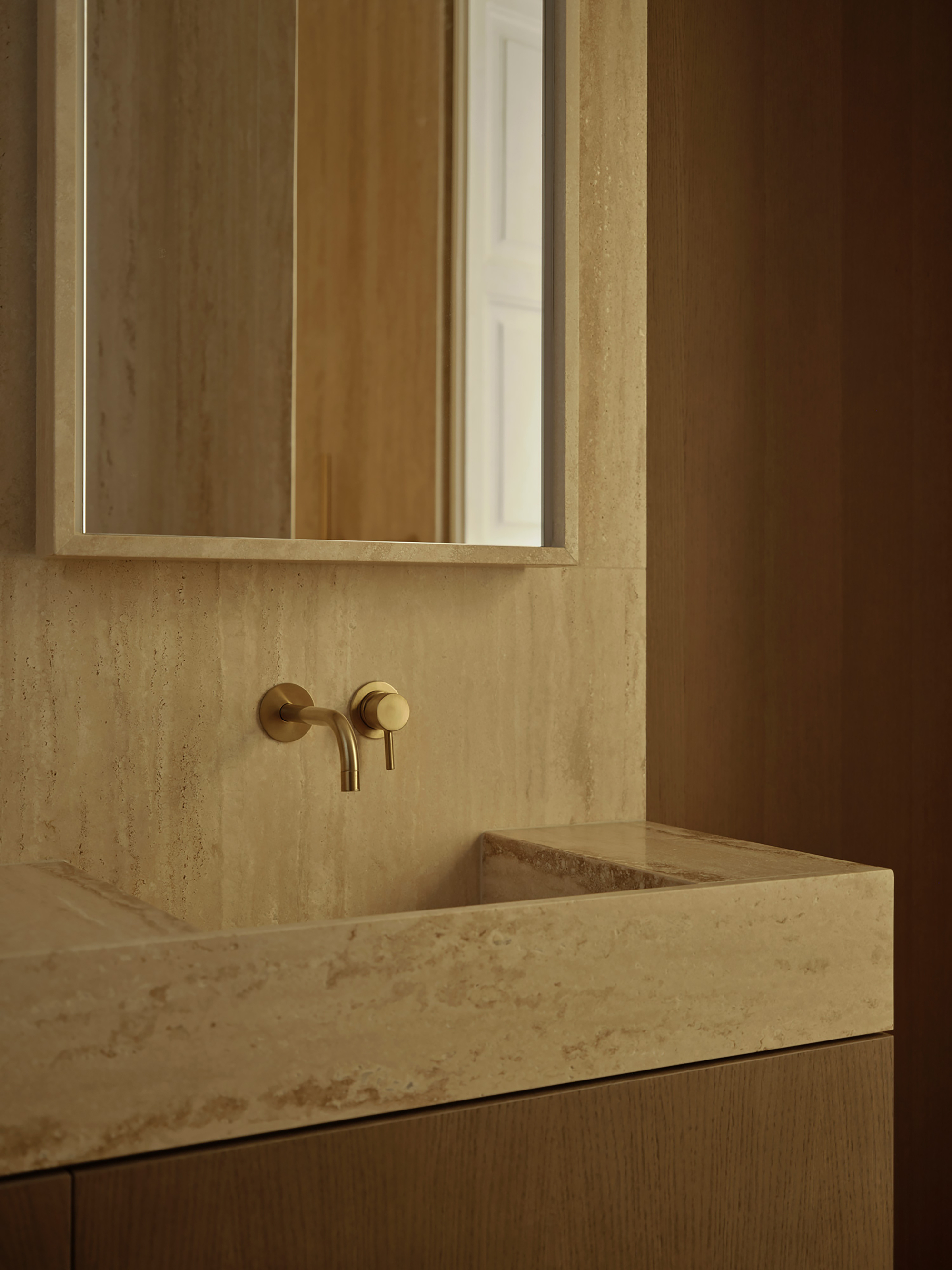 Close-up of a travertine basin with a wall-mounted brass tap and framed mirror, set against warm oak panelling
