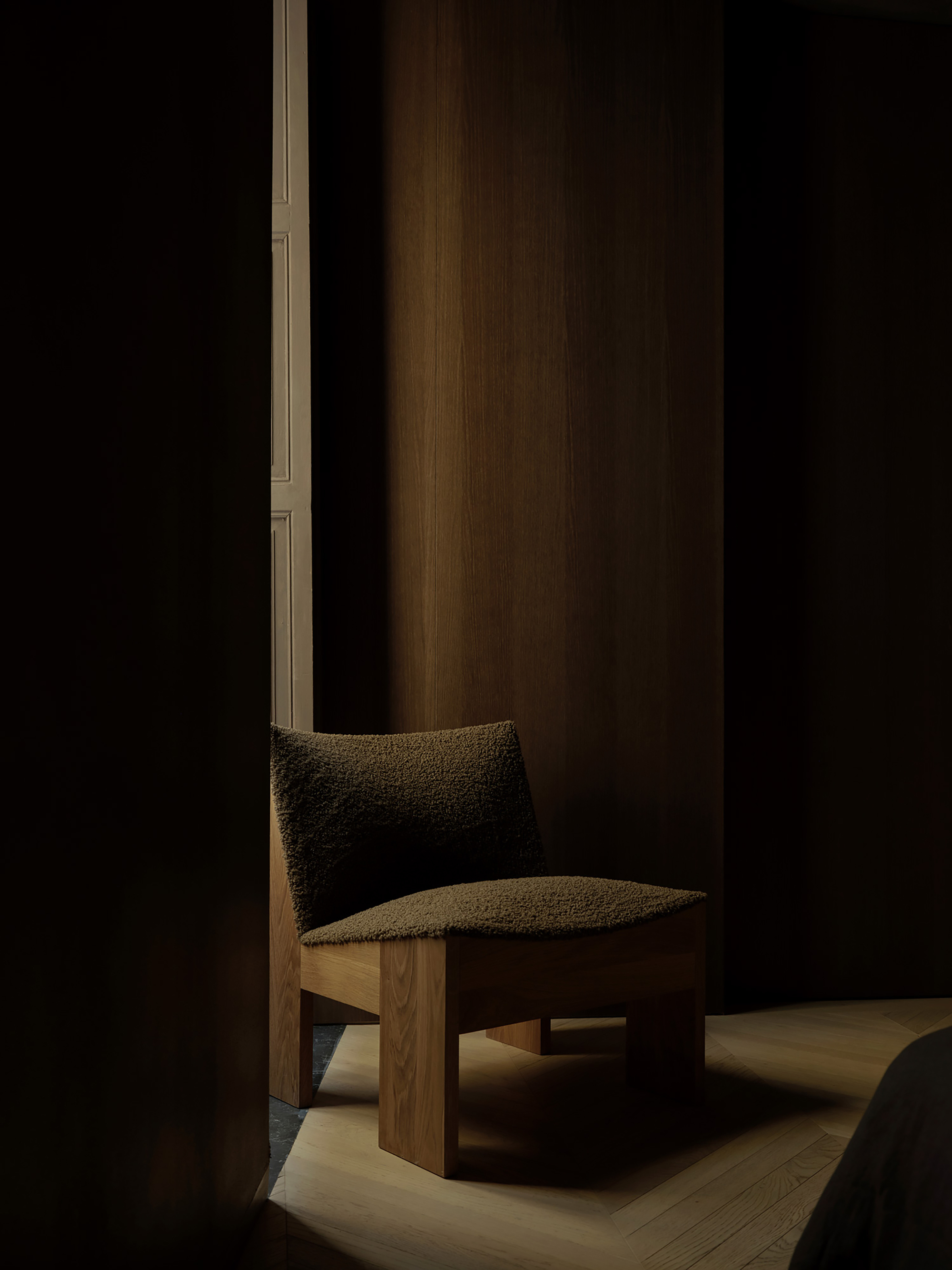 Bouclé lounge chair with a chunky timber base in a dim corner, a sliver of light on the herringbone floor