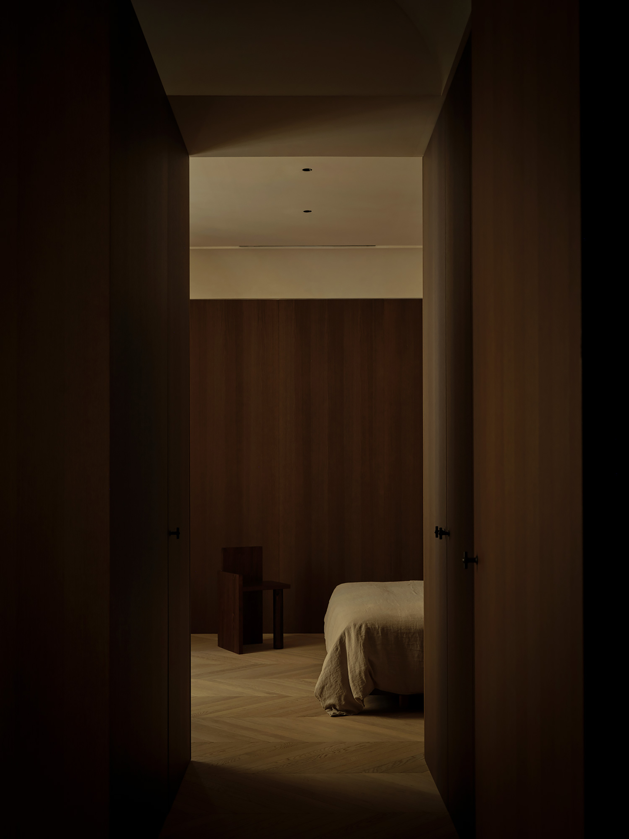 View down a dark timber-clad corridor toward a bed and wooden chair, lit by a soft ceiling glow