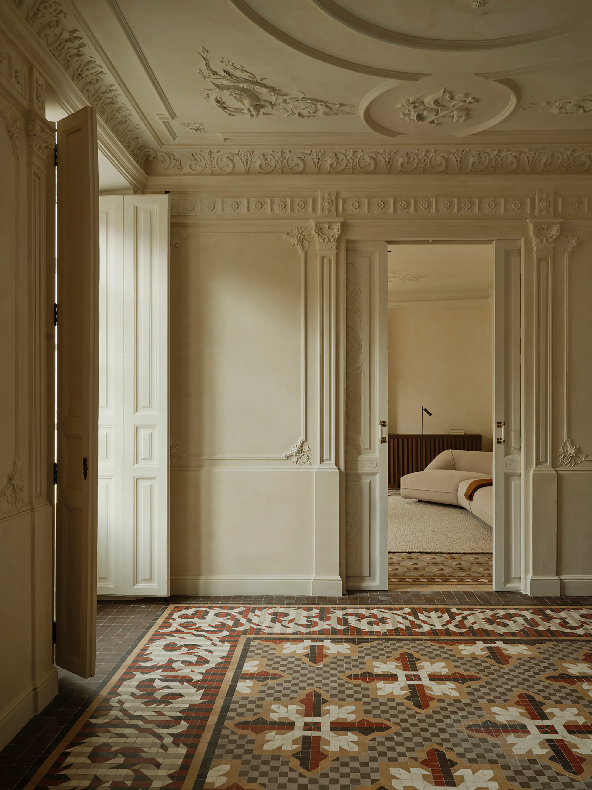 Ornate plaster ceiling and wall mouldings above a patterned tile floor, with a cream sofa glimpsed through the doorway