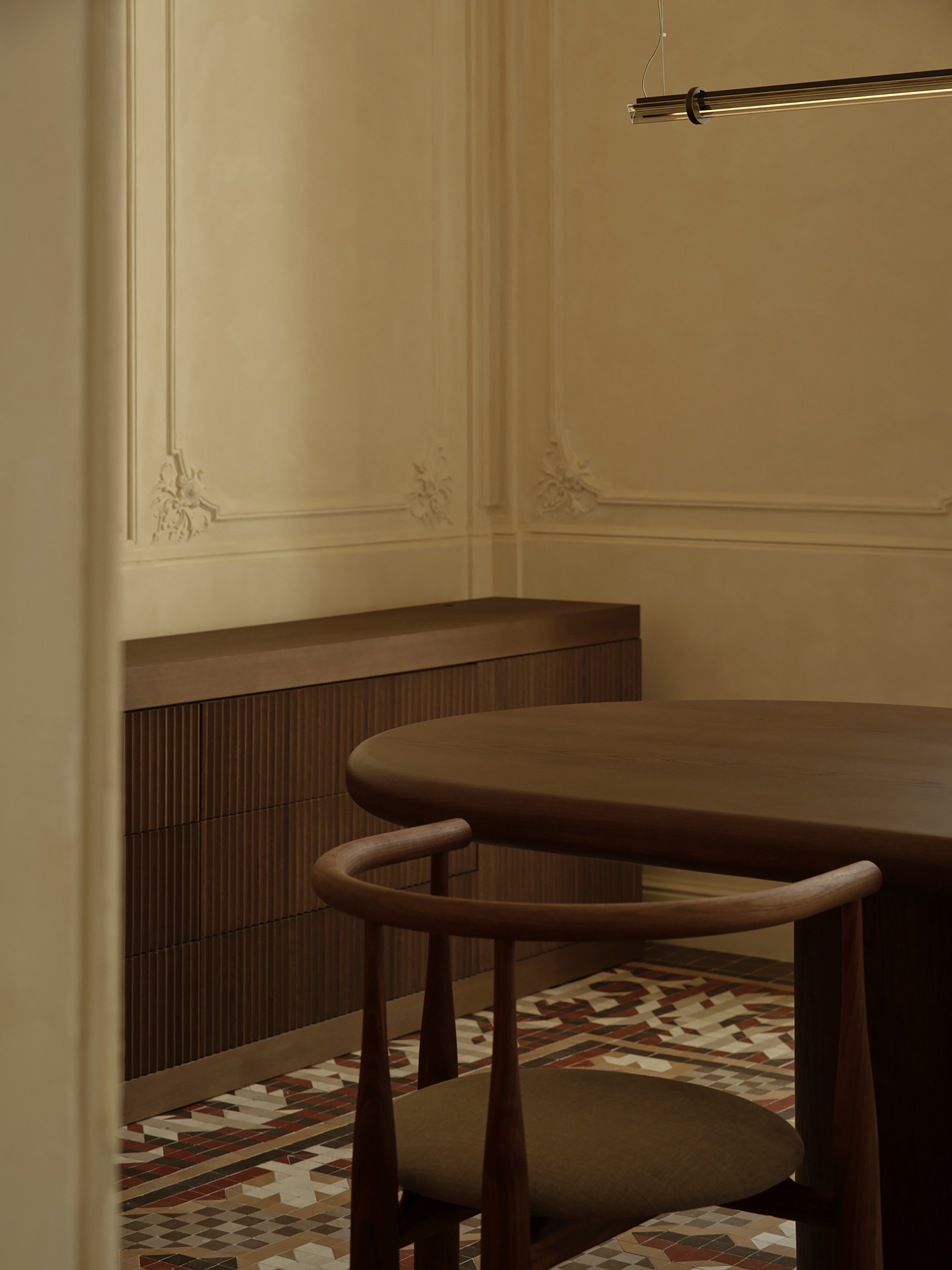 Fluted timber sideboard and rounded walnut table beside a curved-back chair, set against plaster wall moldings and a patterned tile floor in soft light