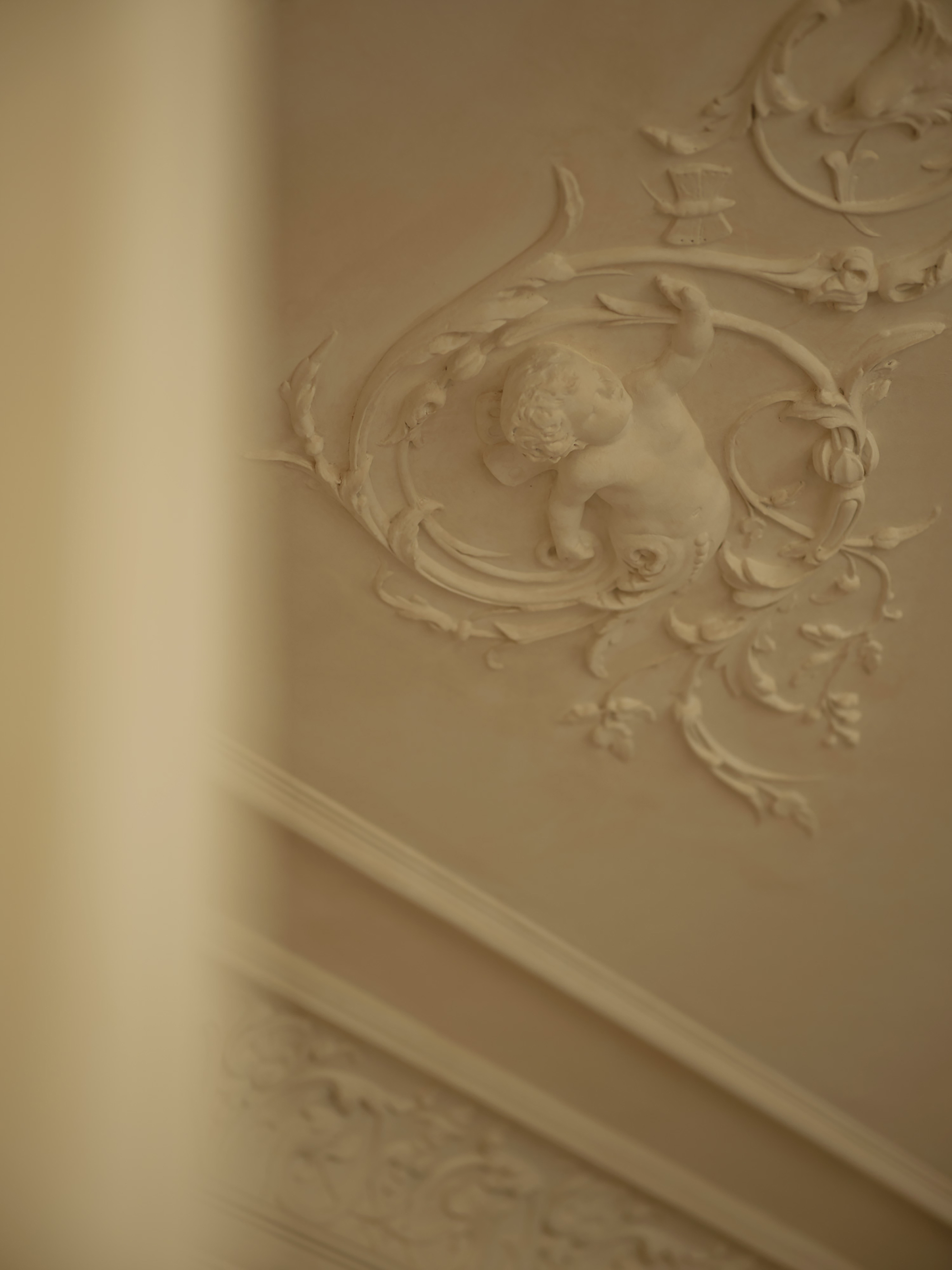 Close-up of a plaster cherub in a scrolling foliate relief on the wall, seen past a blurred column edge