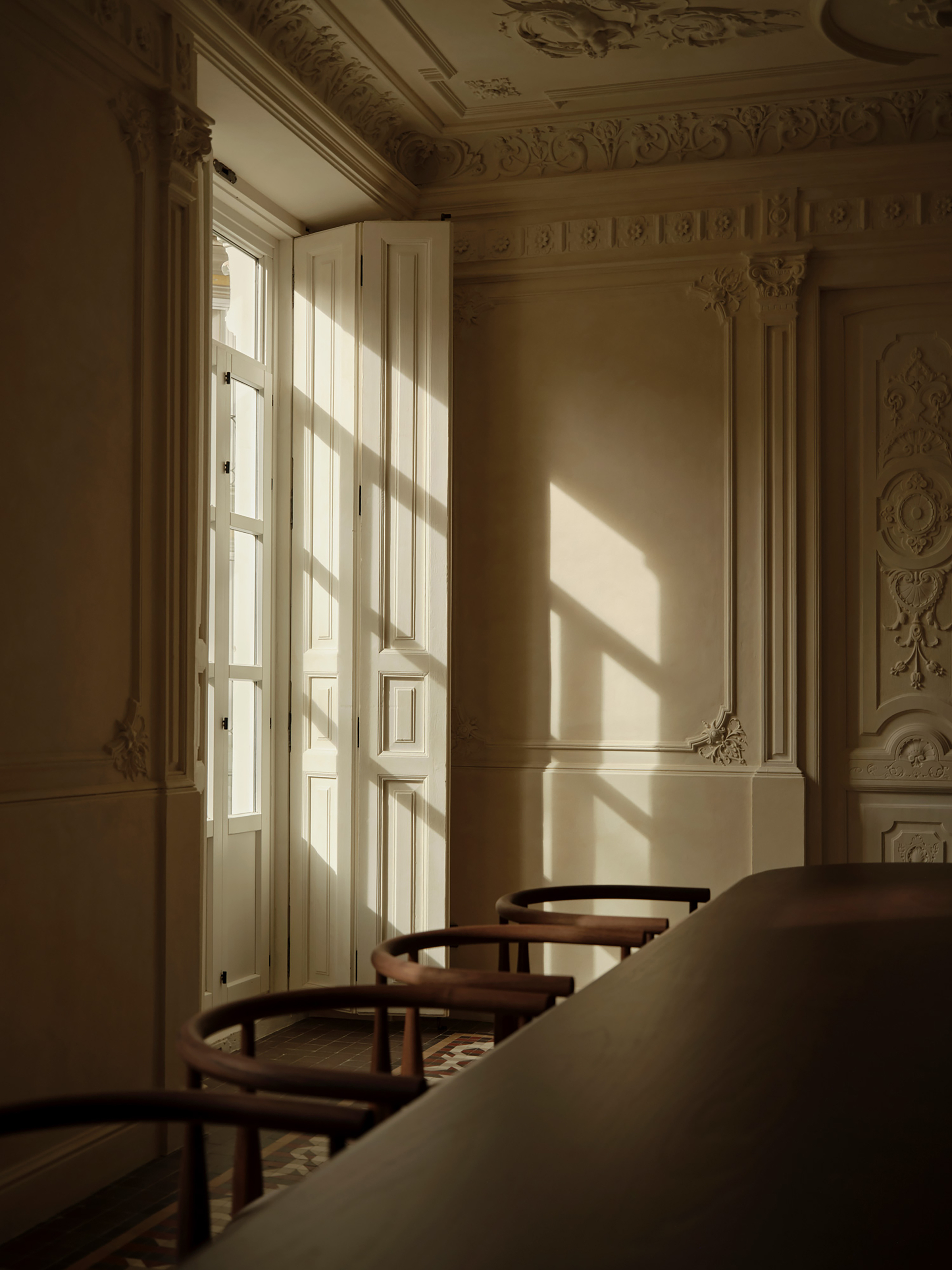 Tall shuttered window casting late sun across ornate plaster walls and a row of curved-back chairs at a long table