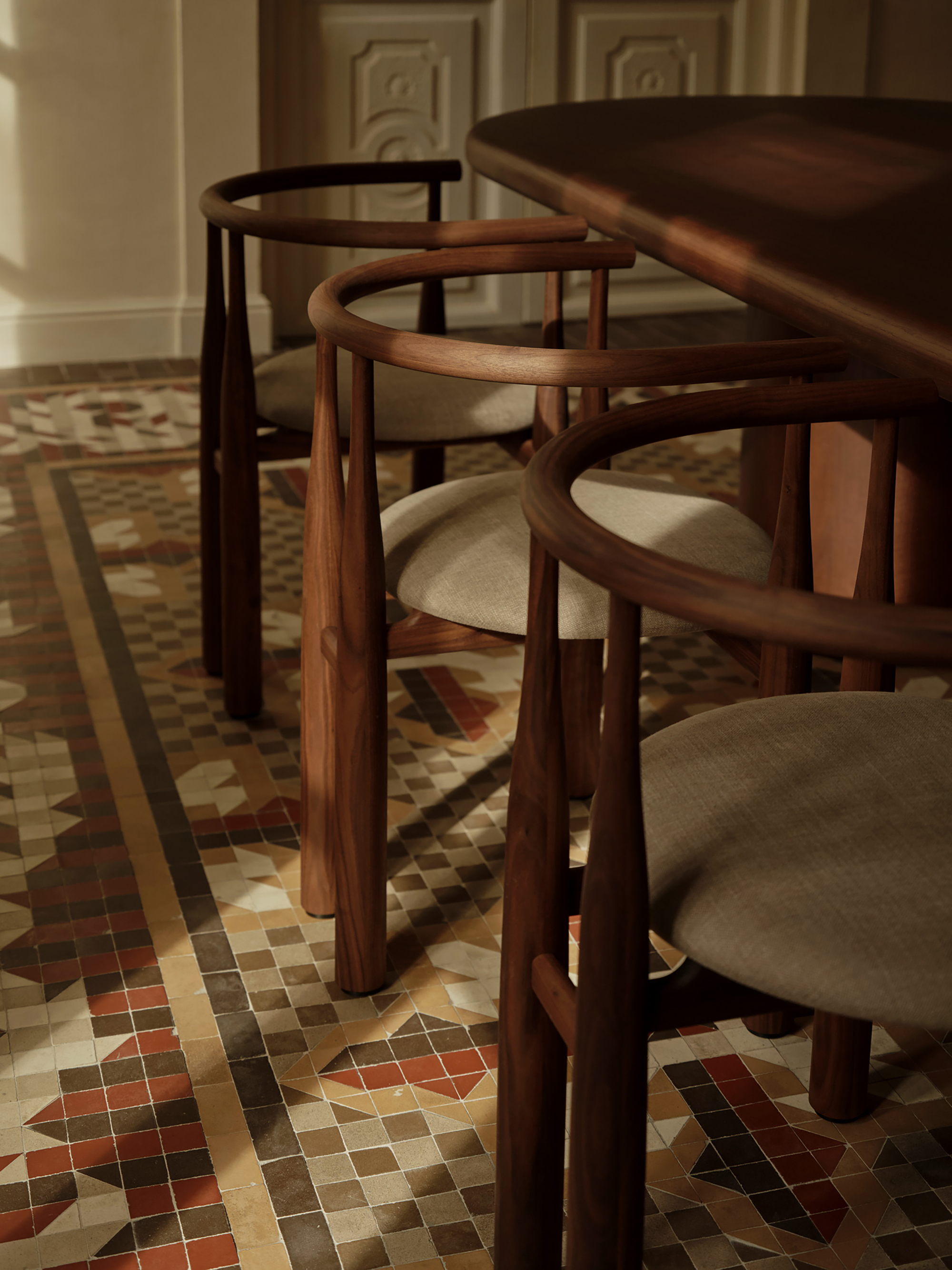 Three curved-back walnut chairs with linen seats on a red and ochre mosaic tile floor in low raking sunlight
