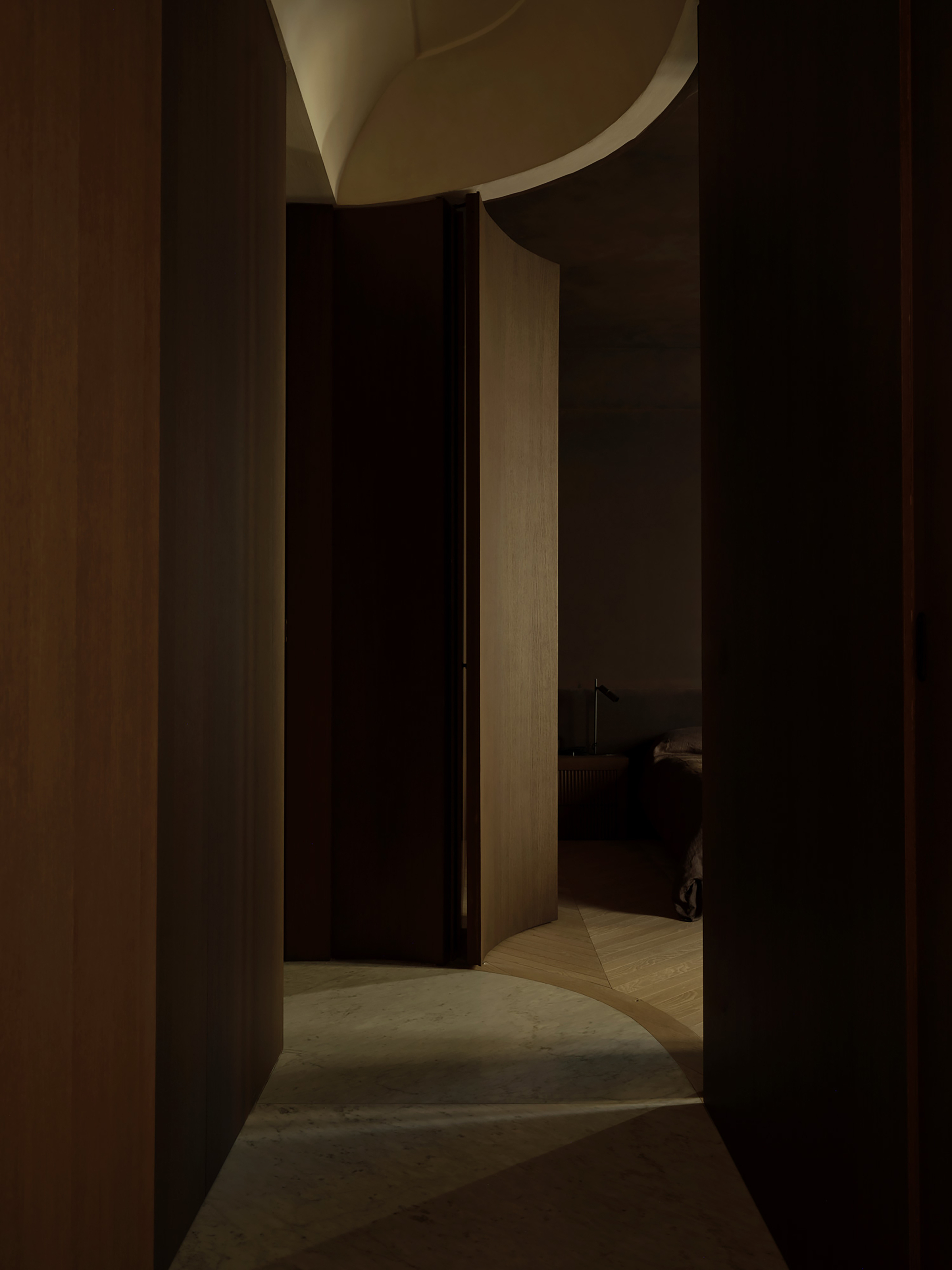 Dim corridor of timber panels curving toward a bedroom, with a marble and herringbone floor edge catching a sliver of light