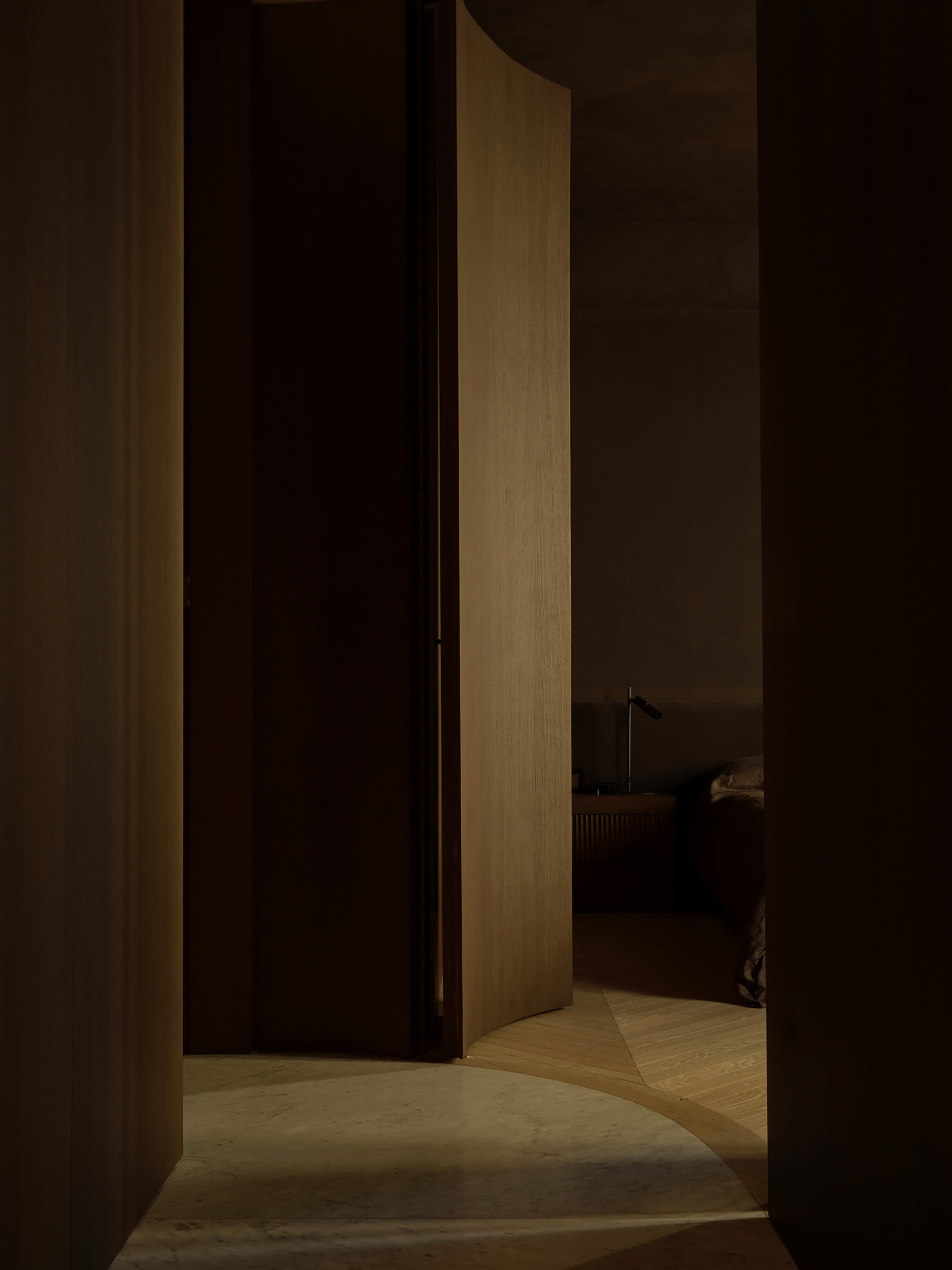 Curved oak door leaf opening onto a dark bedroom with a bedside lamp, floor meeting marble and herringbone parquet in a patch of light