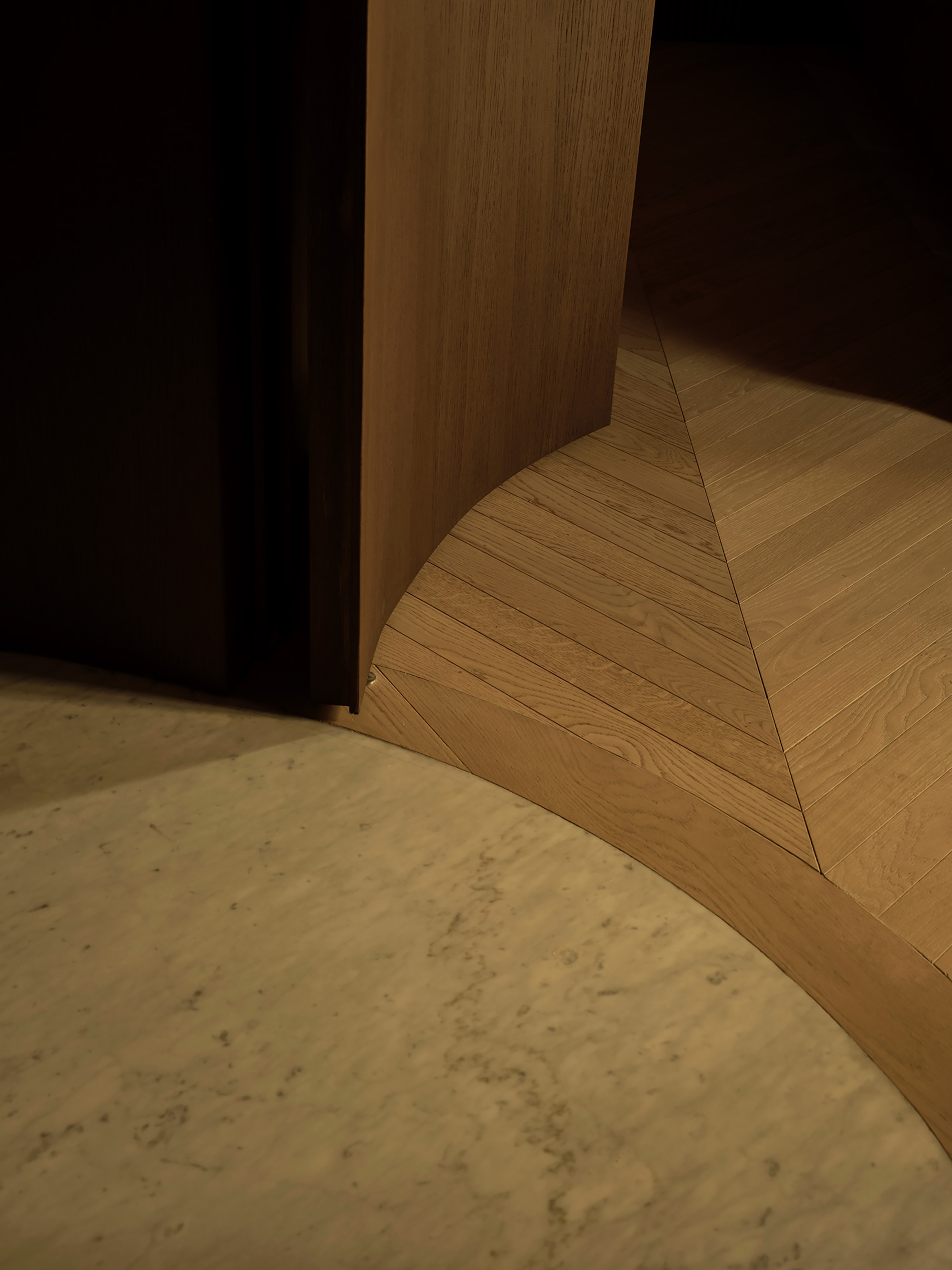 Curved oak door edge meeting a herringbone parquet floor inlaid against a pale marble threshold in low raking light