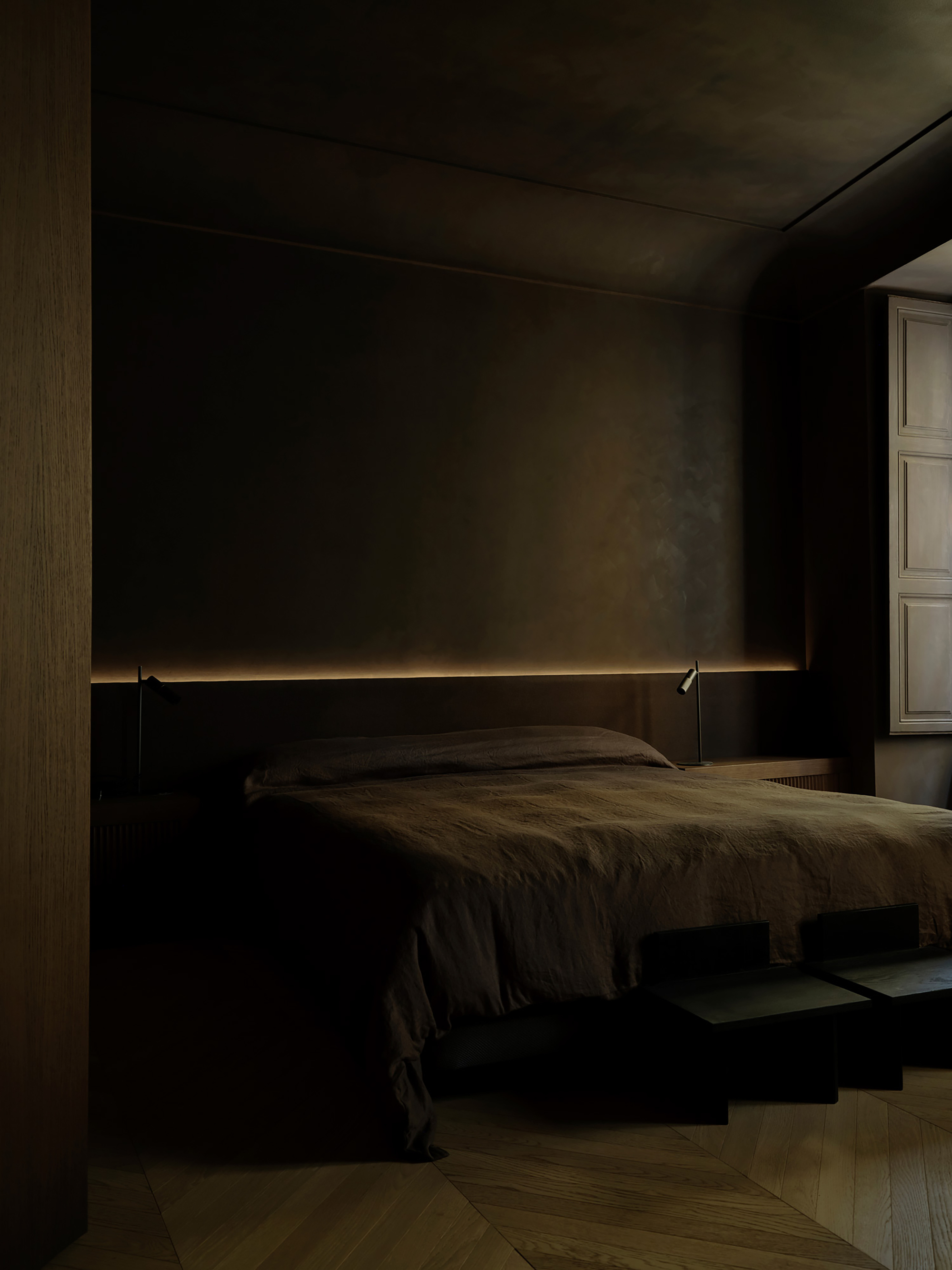 Low bed with rumpled linen under a recessed strip of warm light, with a paneled shutter beside the window