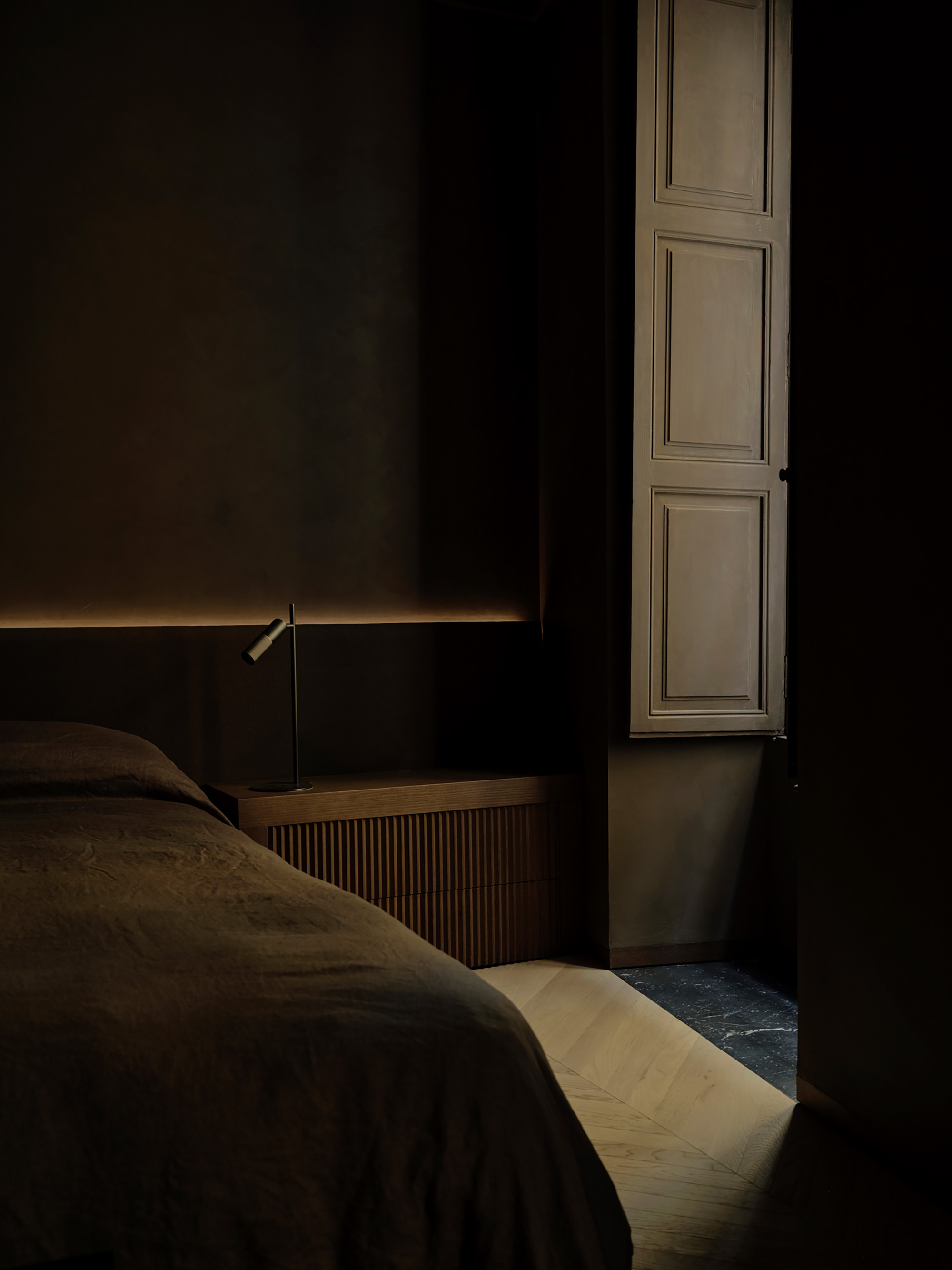 Bedside table with fluted drawer front and a small reading lamp, with a paneled shutter catching daylight