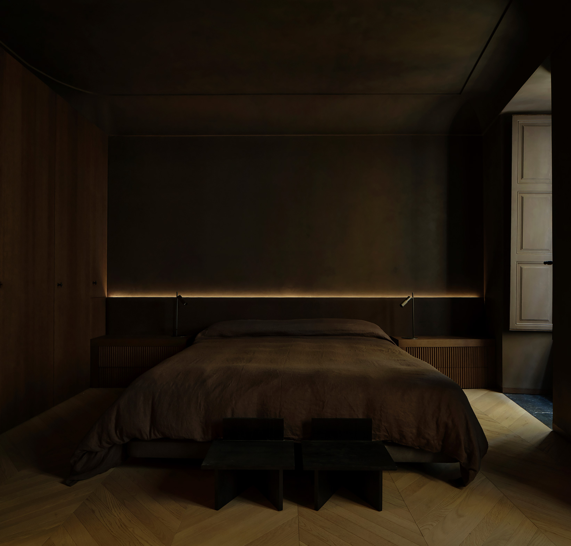 Frontal view of the bed, two dark stools and herringbone floor beneath a softly lit headboard wall