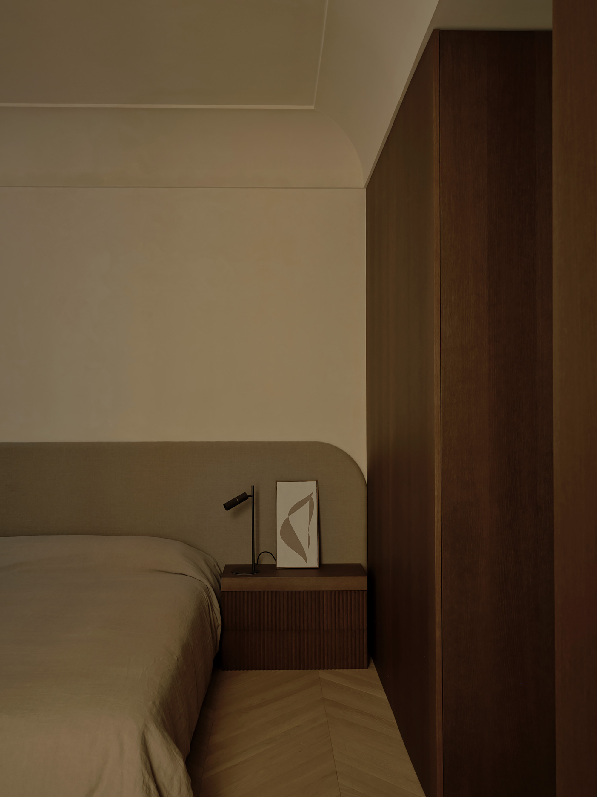 Bedside corner with fluted nightstand, small lamp and framed print beside a tall walnut wardrobe volume in soft daylight