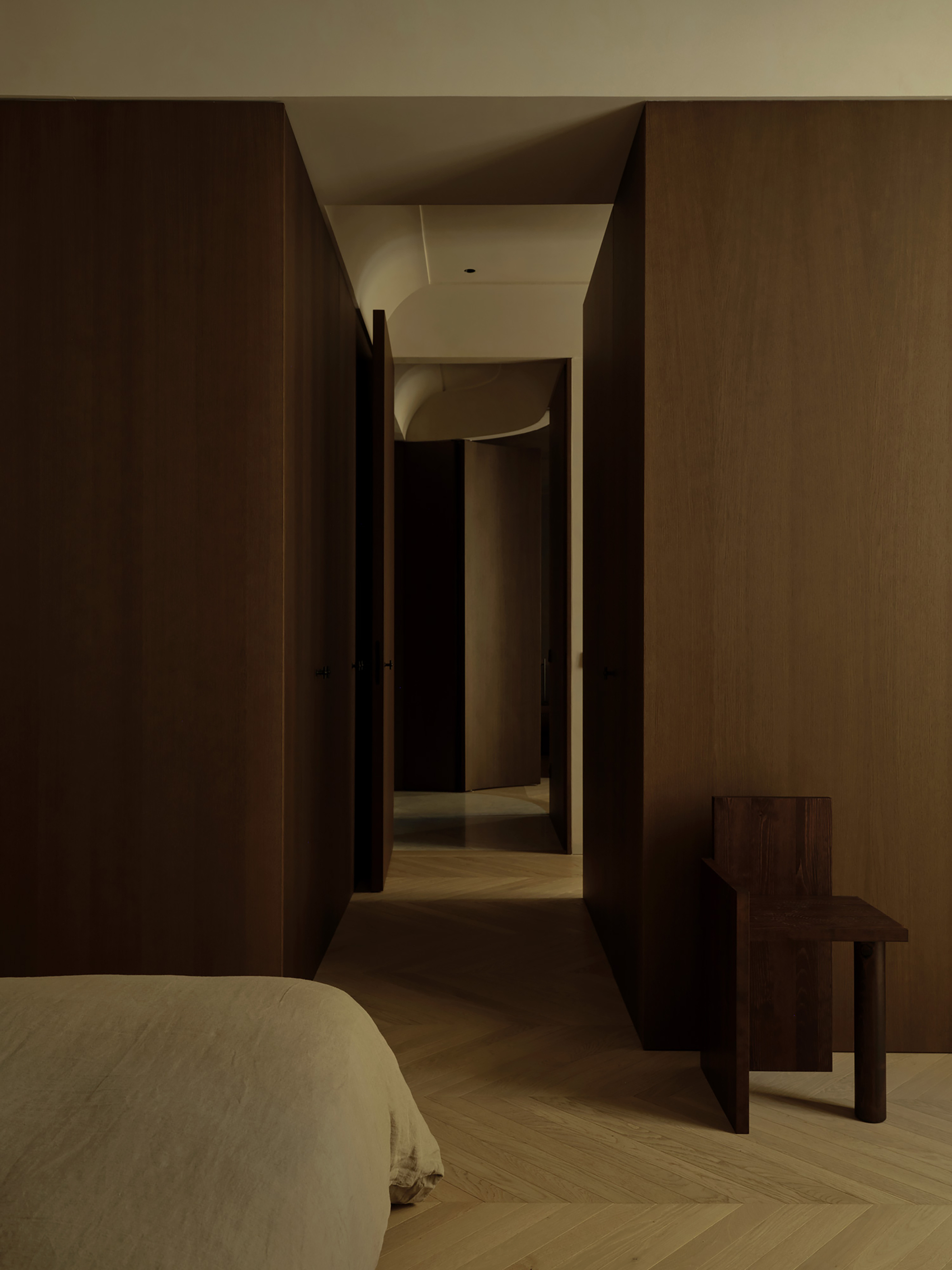 Corridor between dark oak panelled volumes leading past a linen-covered bed and a timber chair on herringbone flooring in dim light