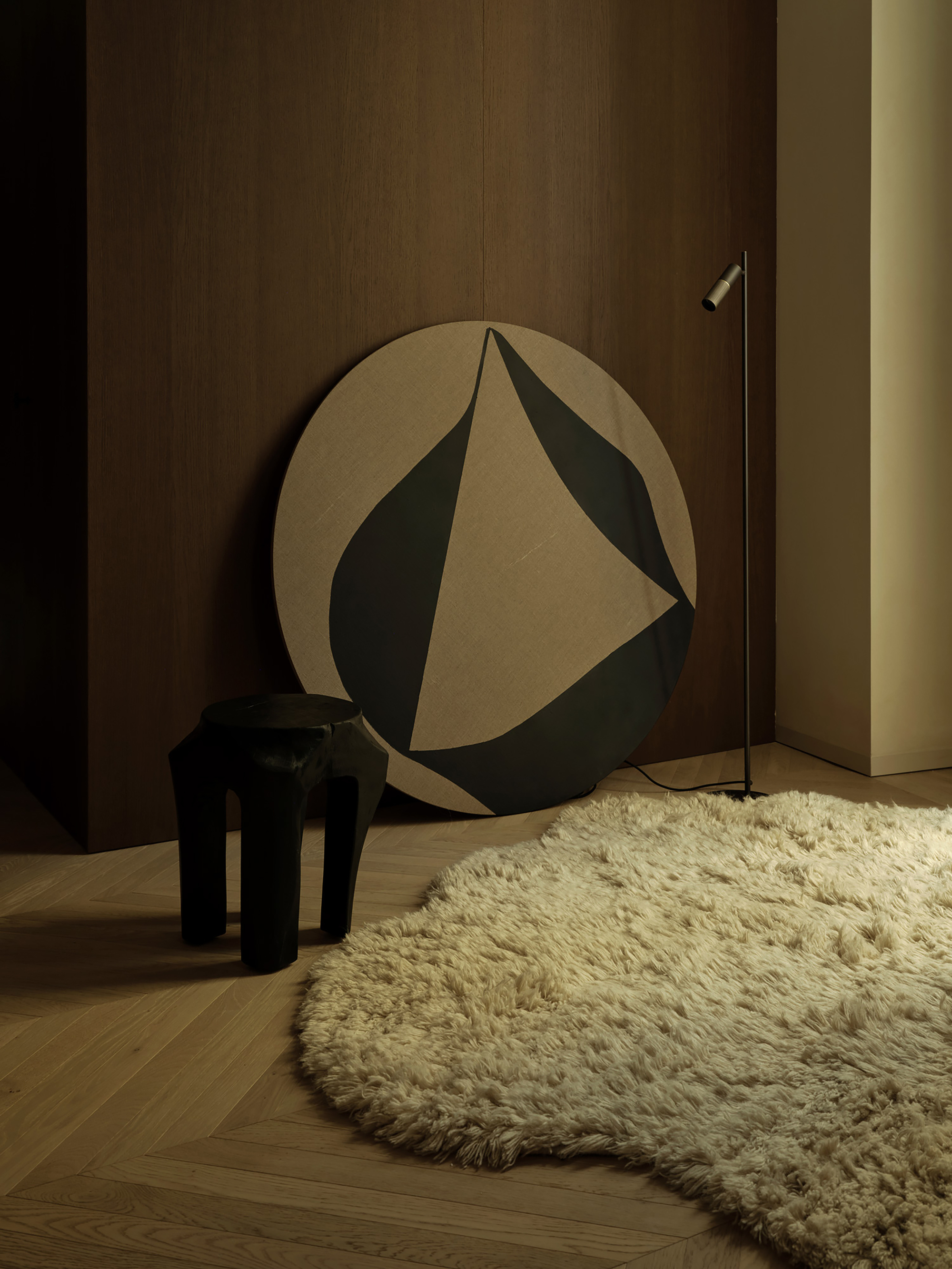 Round canvas with a black leaf-like shape leaning on an oak wall beside a dark stool and shaggy cream rug