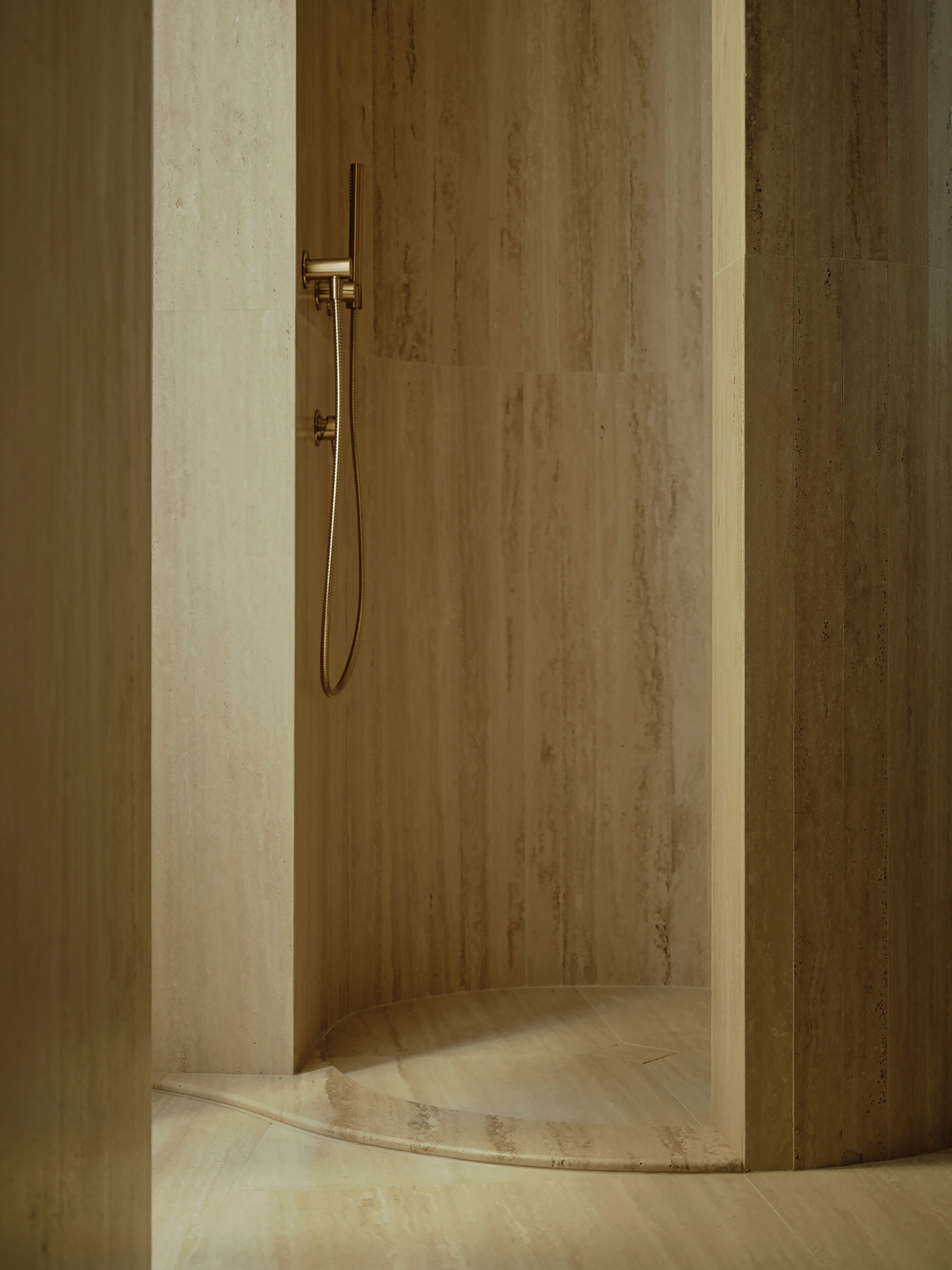 Curved travertine shower alcove with a brass hand shower and a sculpted stone threshold in soft light