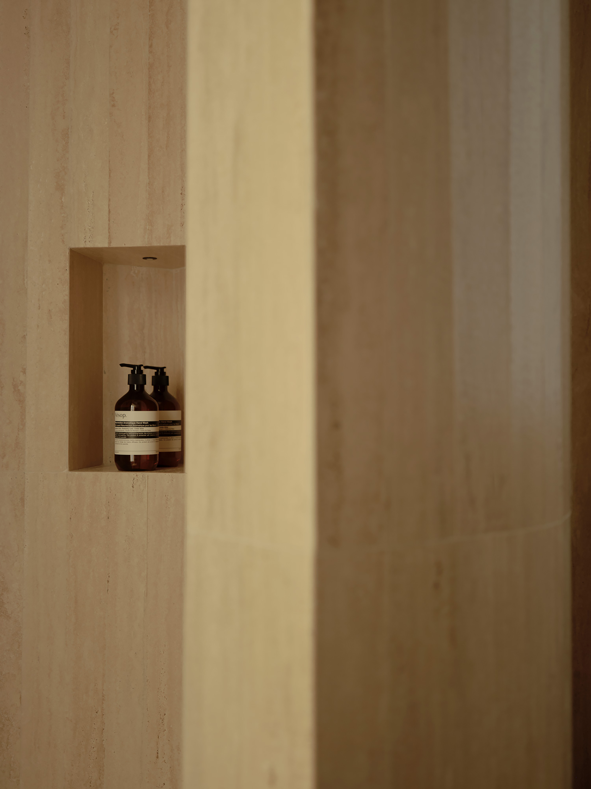 Recessed travertine niche holding two amber pump bottles, framed by a pale stone column edge in warm light